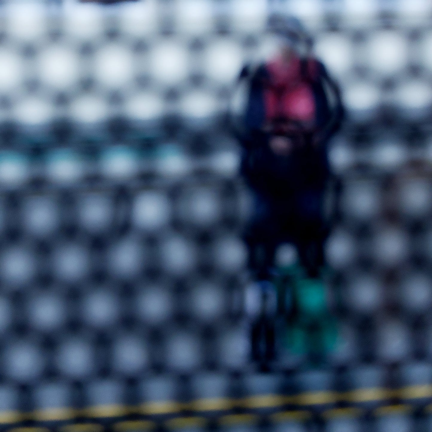 Urban Commute - Series No.2: 9153 - Close Detail View, fine art photograph by Raymond Mather showing a blurred figure in red and navy with a backpack and helmet, standing before a row of colourful train seats. Seen through a circular grid, the image evokes themes of pause, movement, and visual abstraction.
