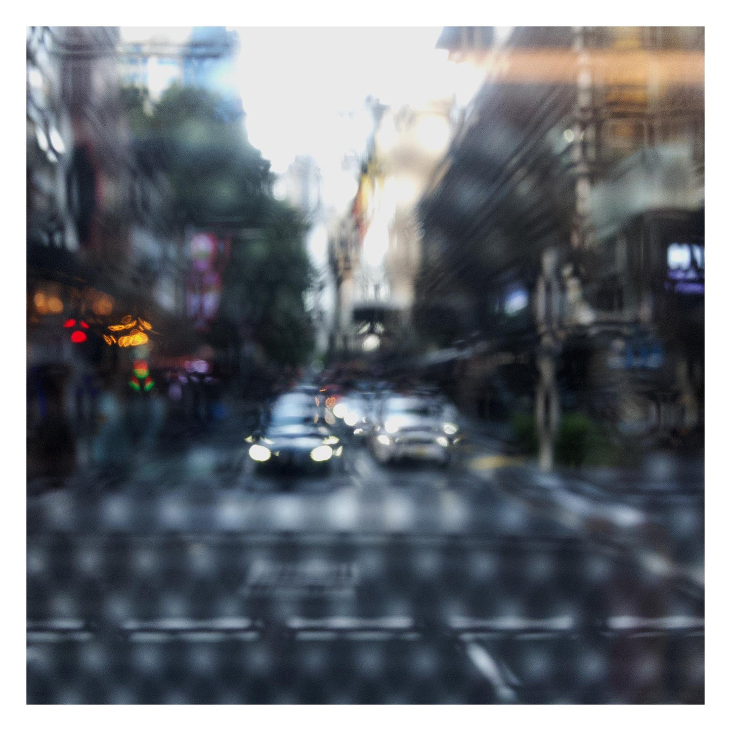 Urban Commute - Series No.2: 9201(1)v1a - 104x104cm, fine art photograph by Raymond Mather showing blurred cars with headlights approaching an intersection, viewed through a patterned surface. City buildings rise on either side, creating a dreamlike abstract of light, rhythm, and perspective.