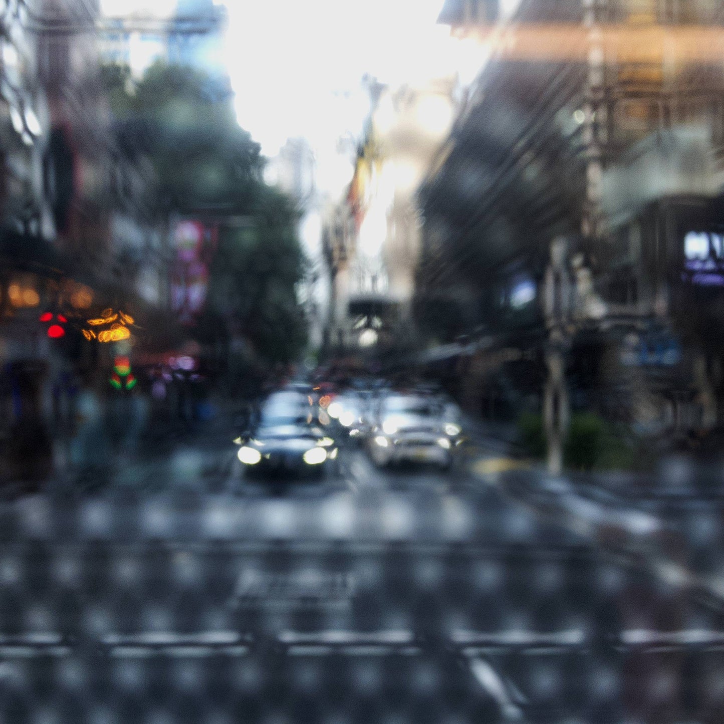 Urban Commute - Series No.2: 9201(1)v1a - 90x90cm, fine art photograph by Raymond Mather showing blurred cars with headlights approaching an intersection, viewed through a patterned surface. City buildings rise on either side, creating a dreamlike abstract of light, rhythm, and perspective.