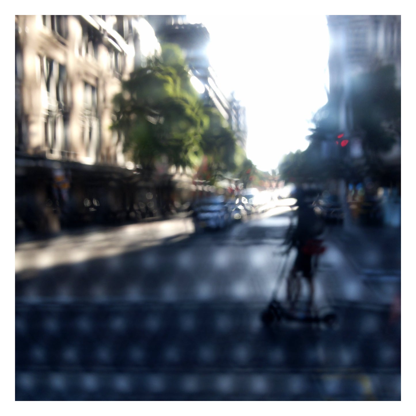 Urban Commute - Series No.2: 9230v1a - 104x104cm, fine art photograph by Raymond Mather featuring a silhouetted scooter rider crossing a sunlit city street. Seen through a patterned screen, the image blends light, blur, and architectural rhythm into a poetic urban abstraction.