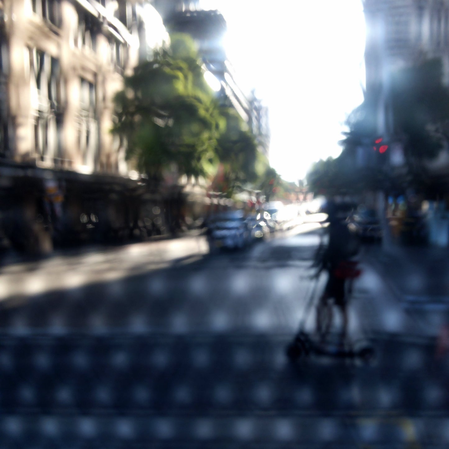 Urban Commute - Series No.2: 9230v1a - 90x90cm, fine art photograph by Raymond Mather featuring a silhouetted scooter rider crossing a sunlit city street. Seen through a patterned screen, the image blends light, blur, and architectural rhythm into a poetic urban abstraction.
