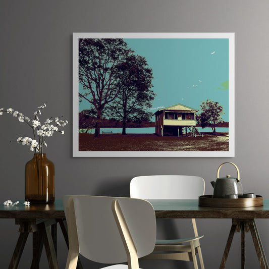Vivid Country - Urunga River House - 6393v44 - Room View, a raised riverfront house stands beside tall silhouetted trees in Urunga, rendered in muted Pop Art tones of teal, brown, and green. A tranquil riverscape with a picnic bench evokes a sense of stillness and simplicity.