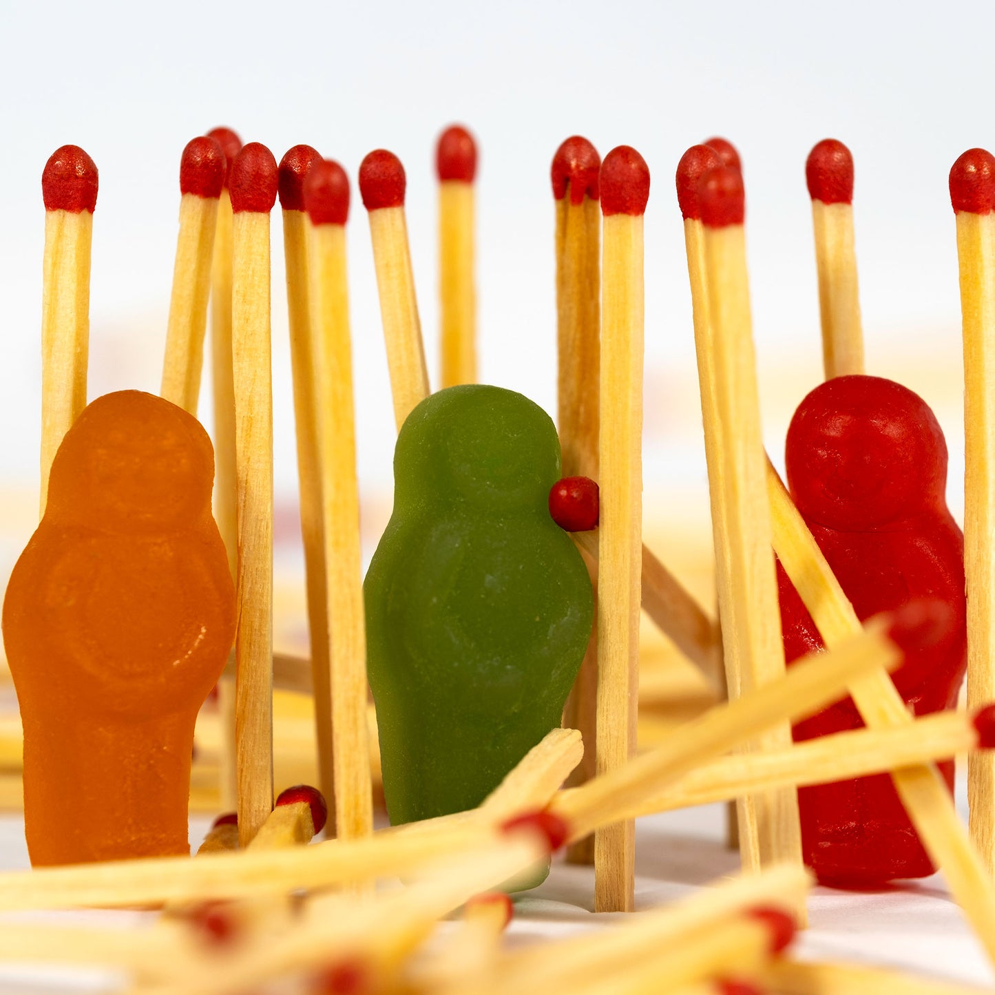 Orange, green and red jelly baby figurines surrounded by upright and scattered red-tipped matches on a white background.