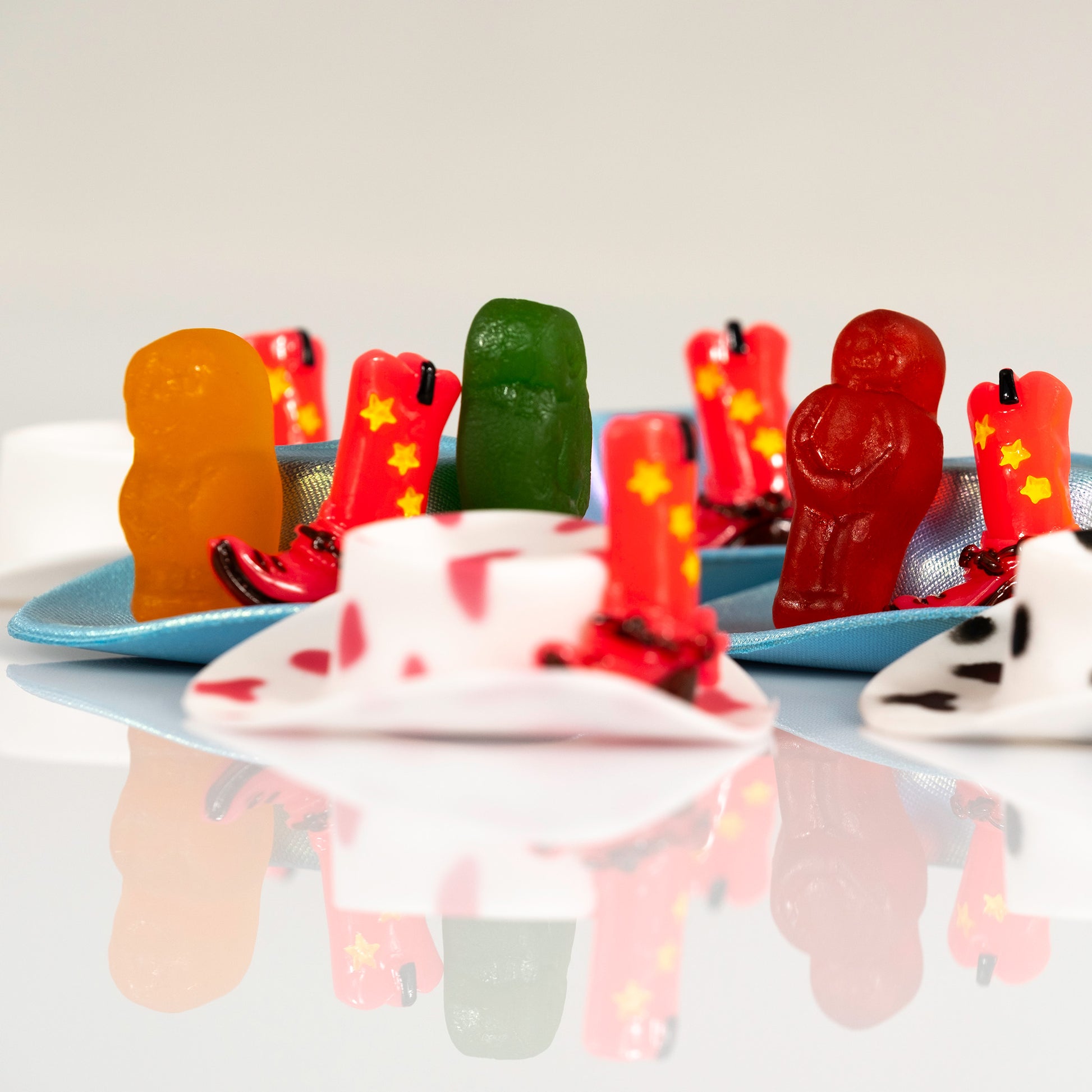 Detail of colourful gummy figures and miniature red cowboy boots arranged with blue hats on a white background.