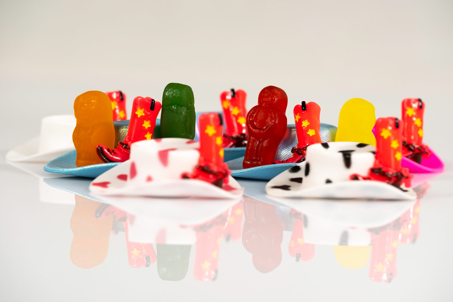 Colourful gummy figures, miniature cowboy boots and sculptural hats reflected on a glossy white surface.