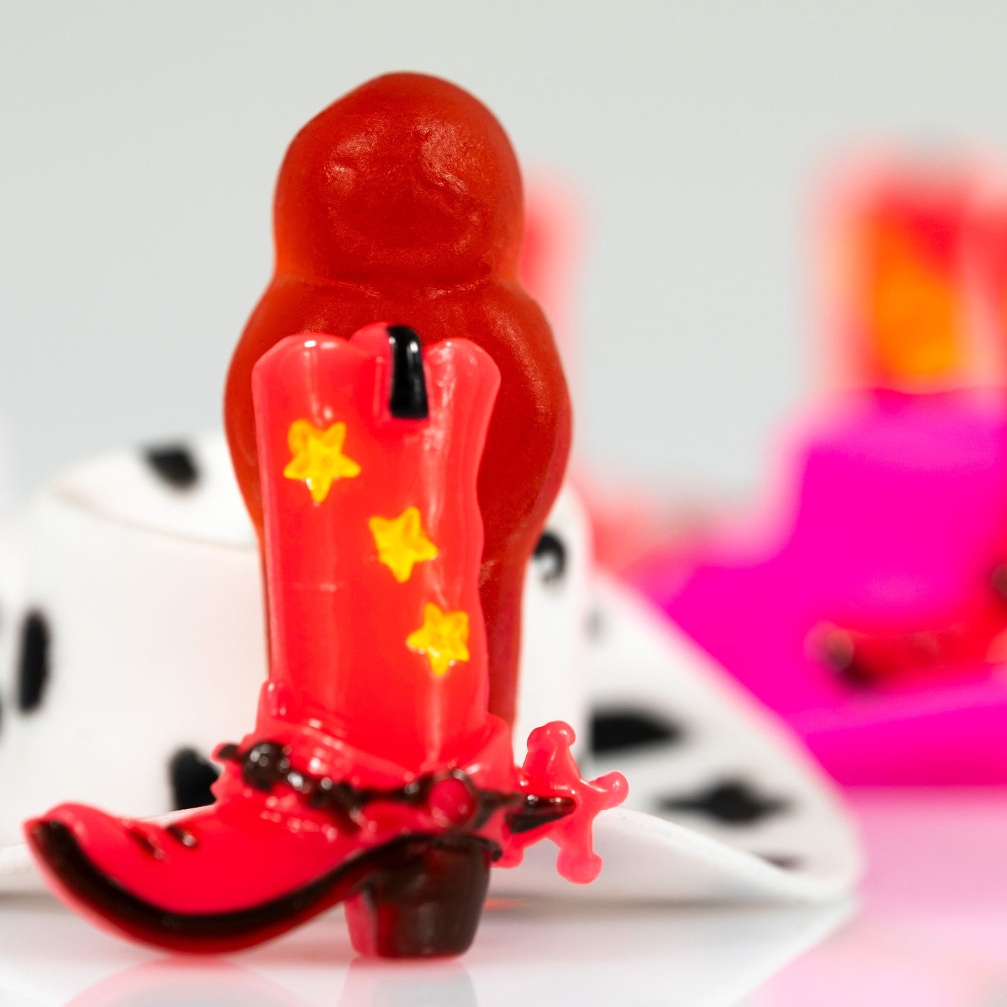 Close crop of Boot Scootin’ Baby 266831v1 featuring a miniature red cowboy boot with yellow stars and a red jelly baby against vivid pink, black and white forms.
