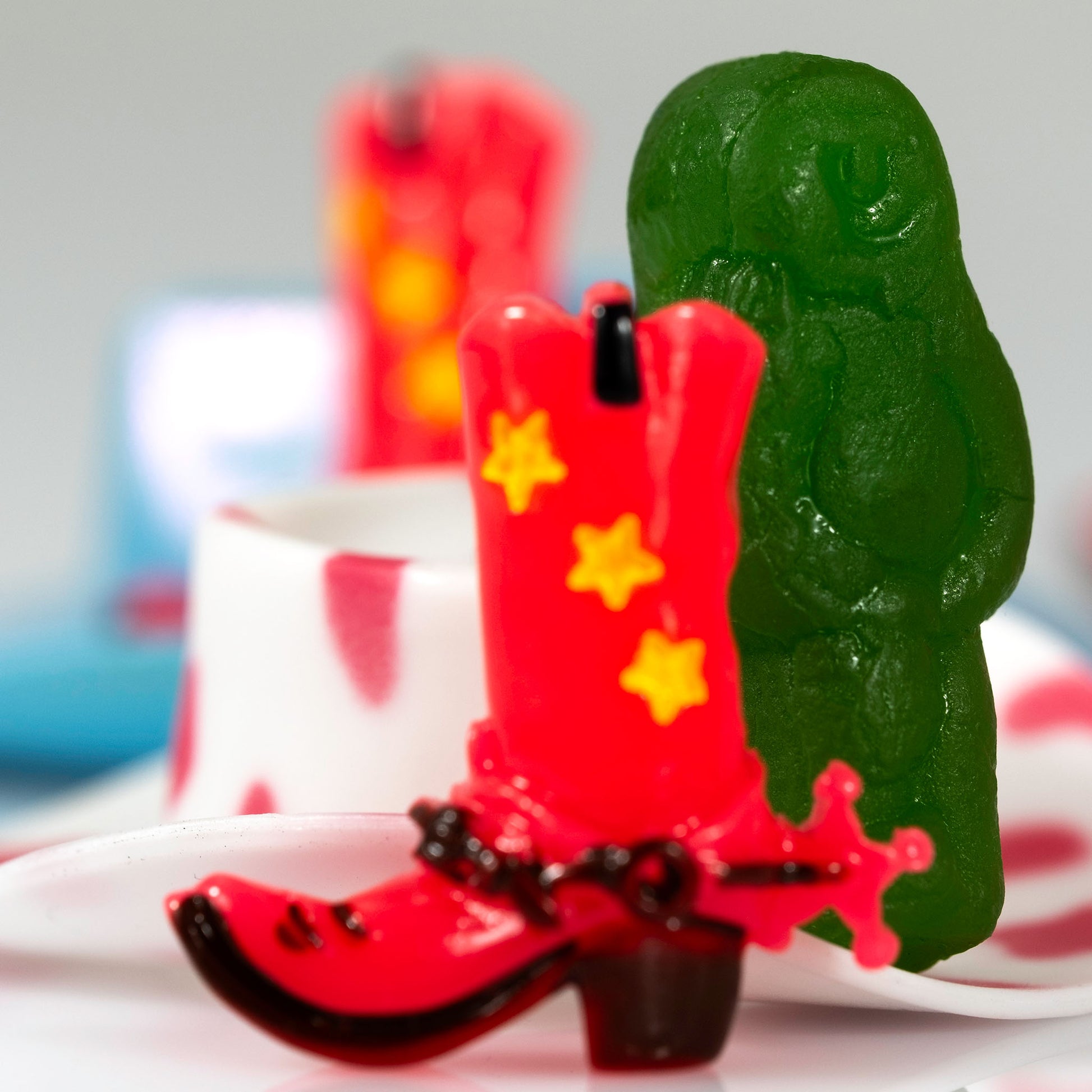 Close crop of Boot Scootin’ Baby 266835v1 featuring a bright red miniature cowboy boot and large green jelly baby against softly blurred colourful objects.