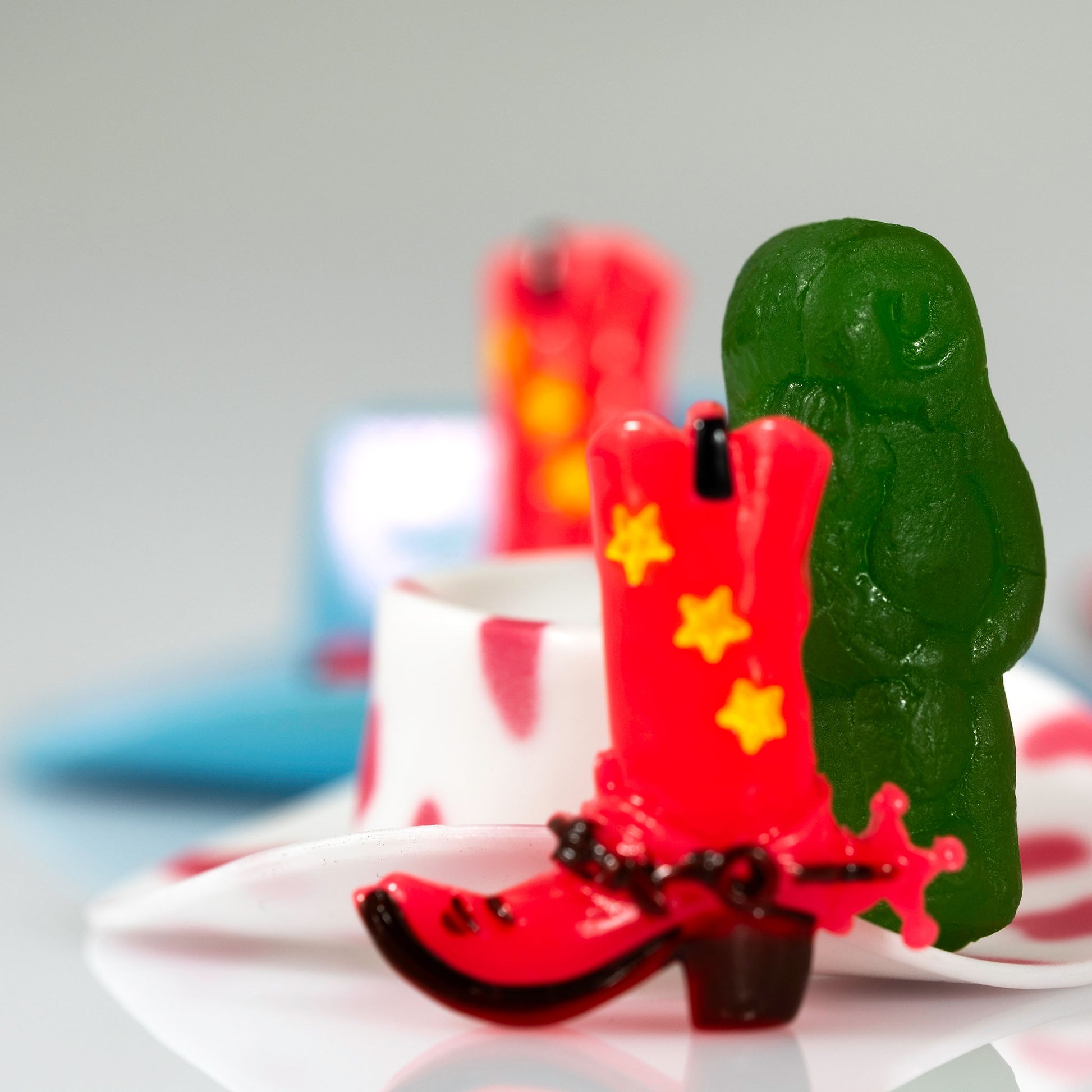 Boot Scootin’ Baby 266835v1, a square contemporary still life photograph of a red cowboy boot positioned in front of a green jelly baby with red, white and blue background elements.