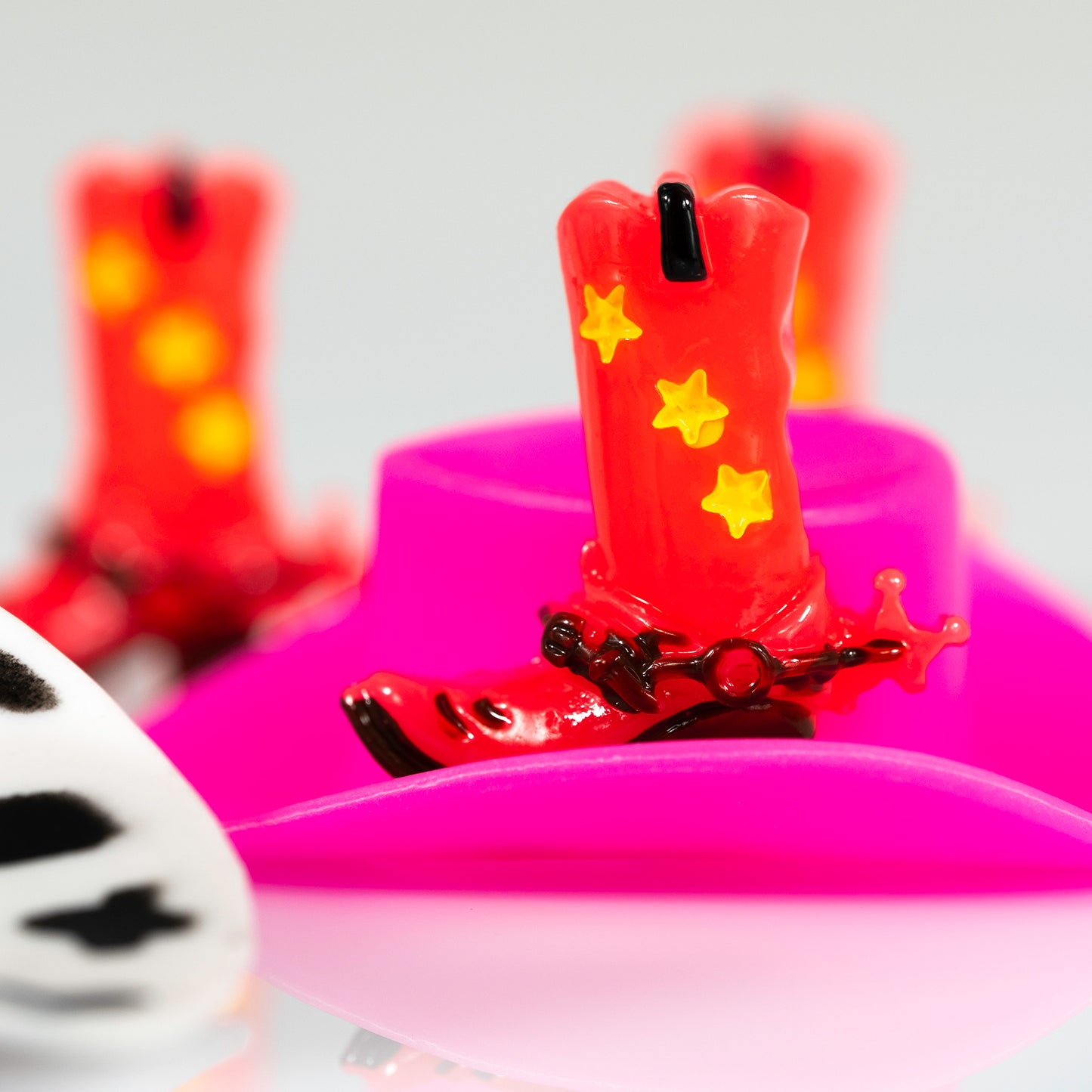 Boot Scootin’ Baby 266837v1, a square contemporary still life photograph of a red cowboy boot with yellow stars against hot pink forms, repeated boots and a black-and-white patterned object.