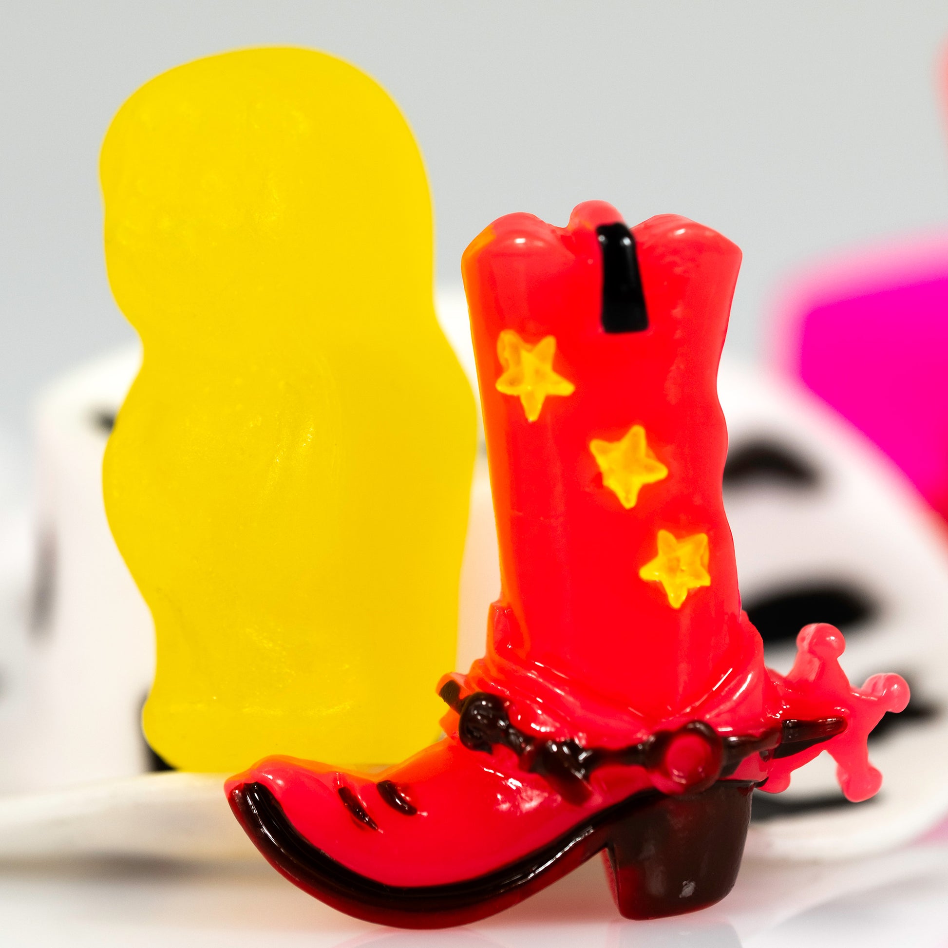 Close crop of Boot Scootin’ Baby 266838v1 featuring a glossy miniature red cowboy boot with yellow stars beside a large luminous yellow figure against a softly blurred background.