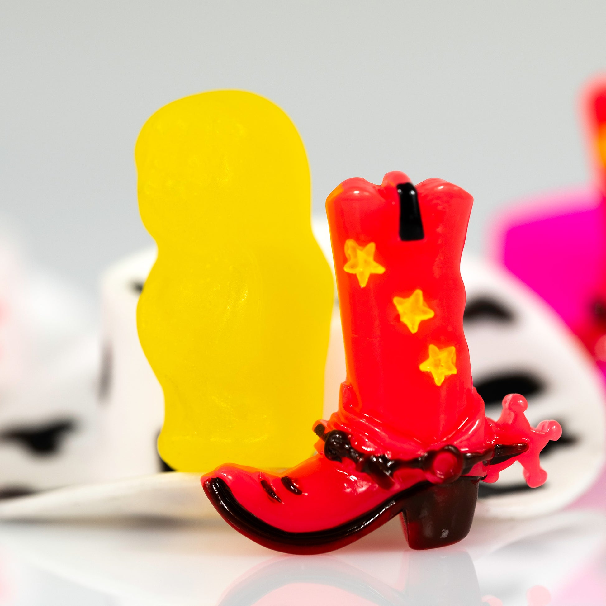 Boot Scootin’ Baby 266838v1, a square contemporary still life photograph of a red cowboy boot beside a vivid yellow figure with black-and-white and hot pink forms in the background.