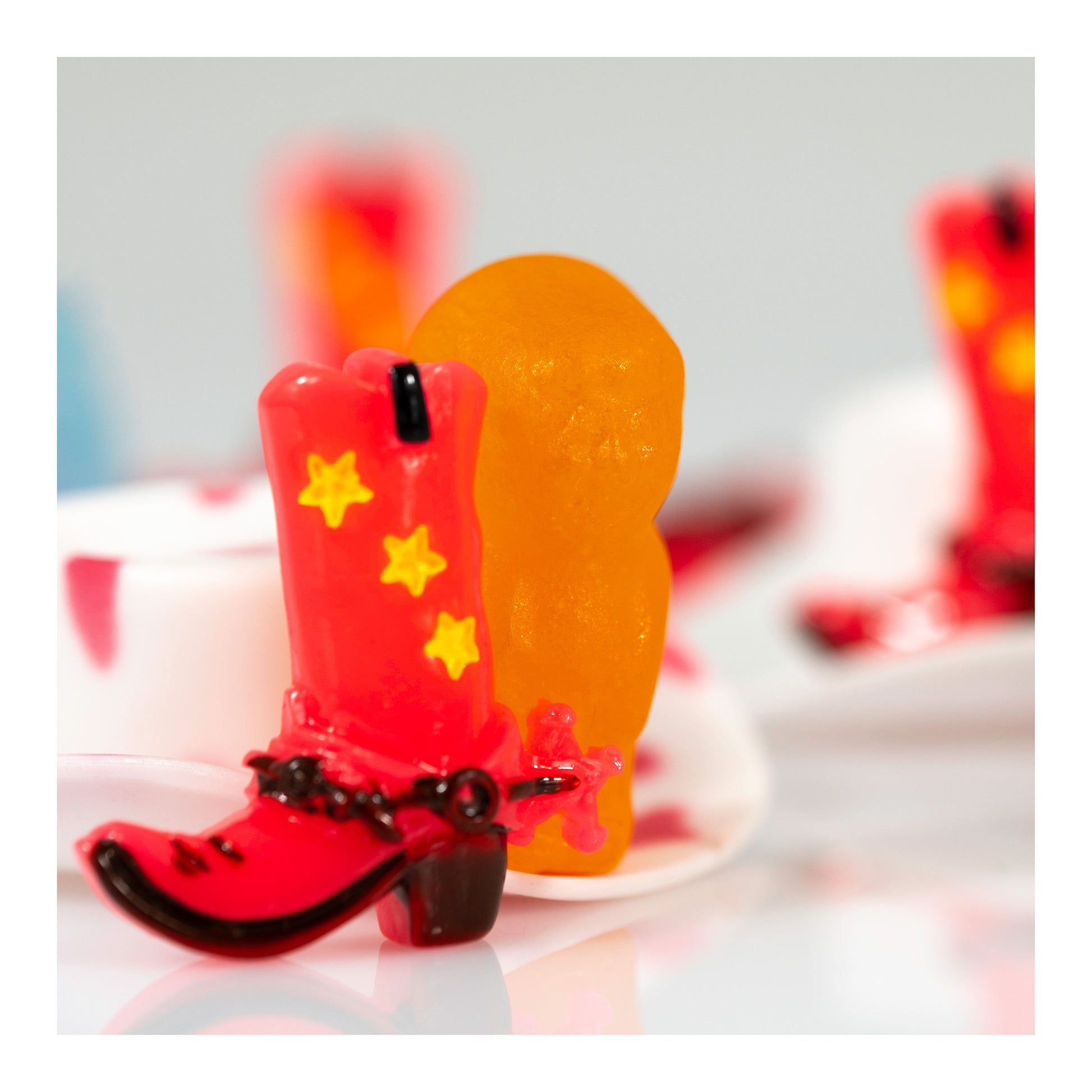 Boot Scootin’ Baby 266841v1 showing a miniature red cowboy boot with yellow stars beside a glowing orange figure, with softly blurred red and white objects behind.