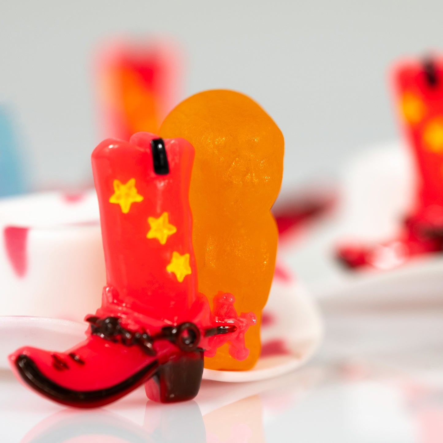 Square contemporary still life photograph of a red cowboy boot and orange figure surrounded by softly focused red, white and blue miniature objects.