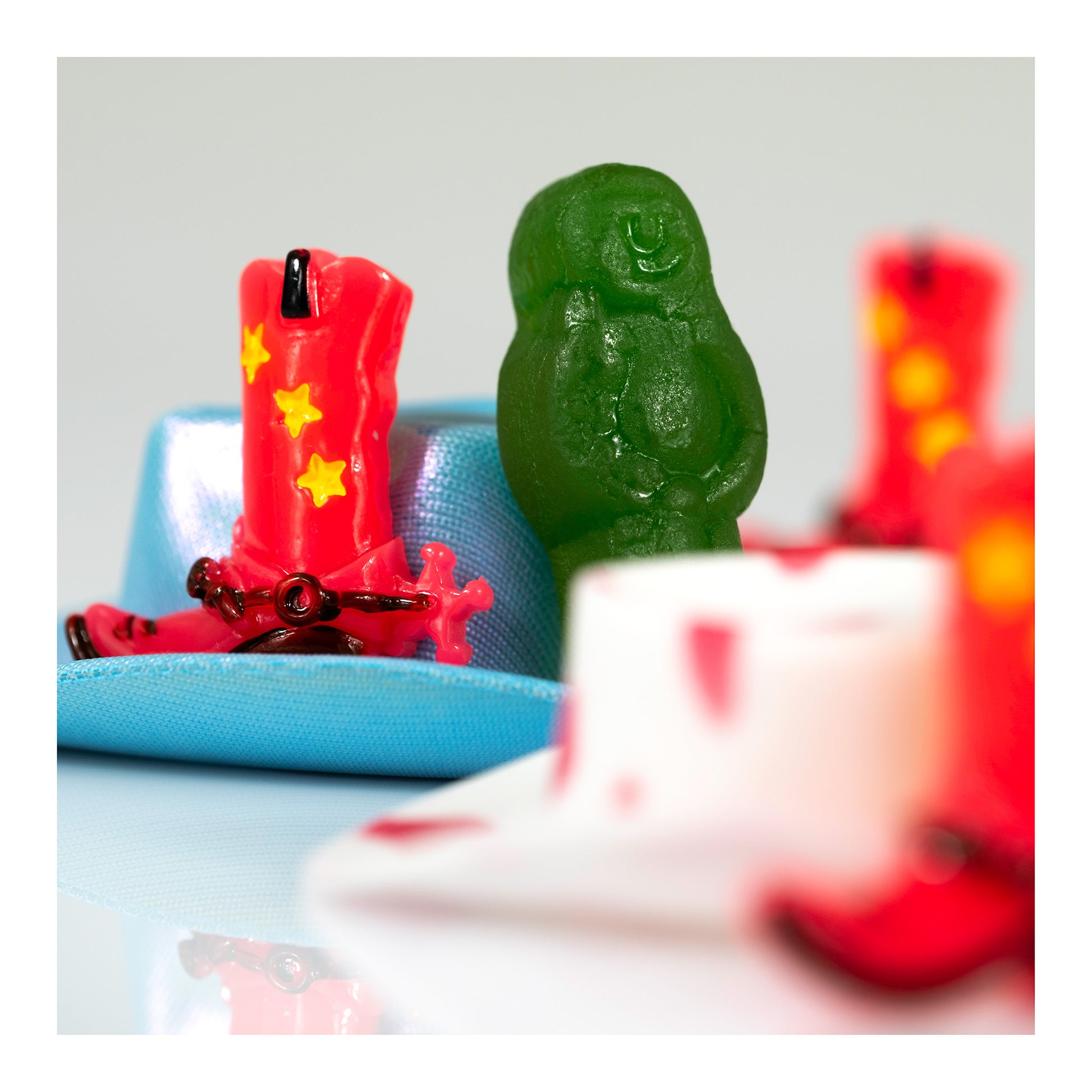 Red miniature cowboy boot beside a green figure on a turquoise platform, surrounded by softly focused red and white objects.