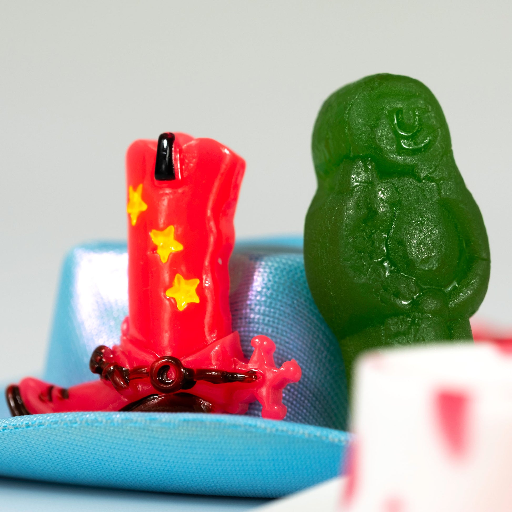Close view of a miniature red cowboy boot with yellow stars beside a green figure, with turquoise and white objects creating layered depth.
