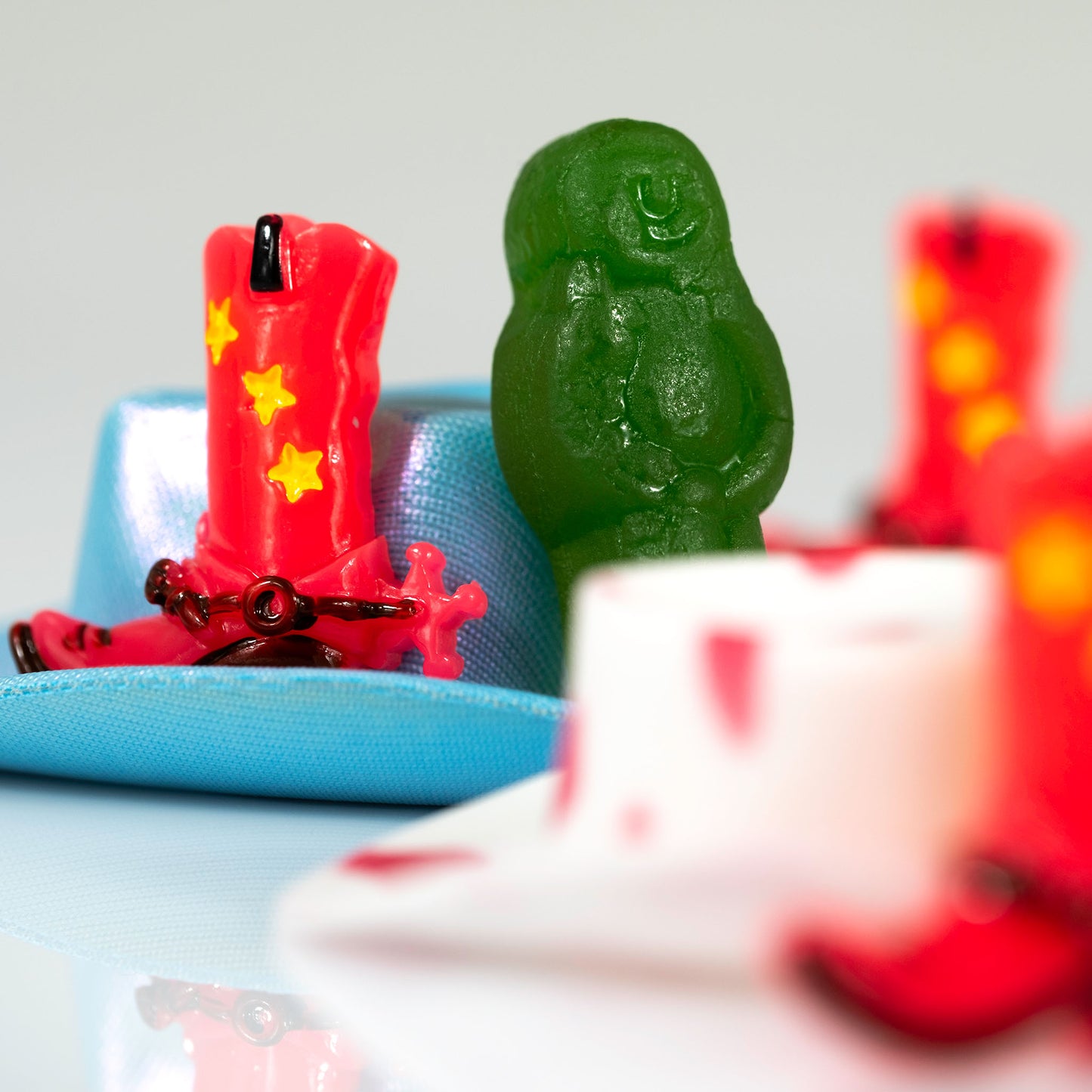 Square still life photograph of a red cowboy boot and green figure with turquoise, white and blurred red objects.