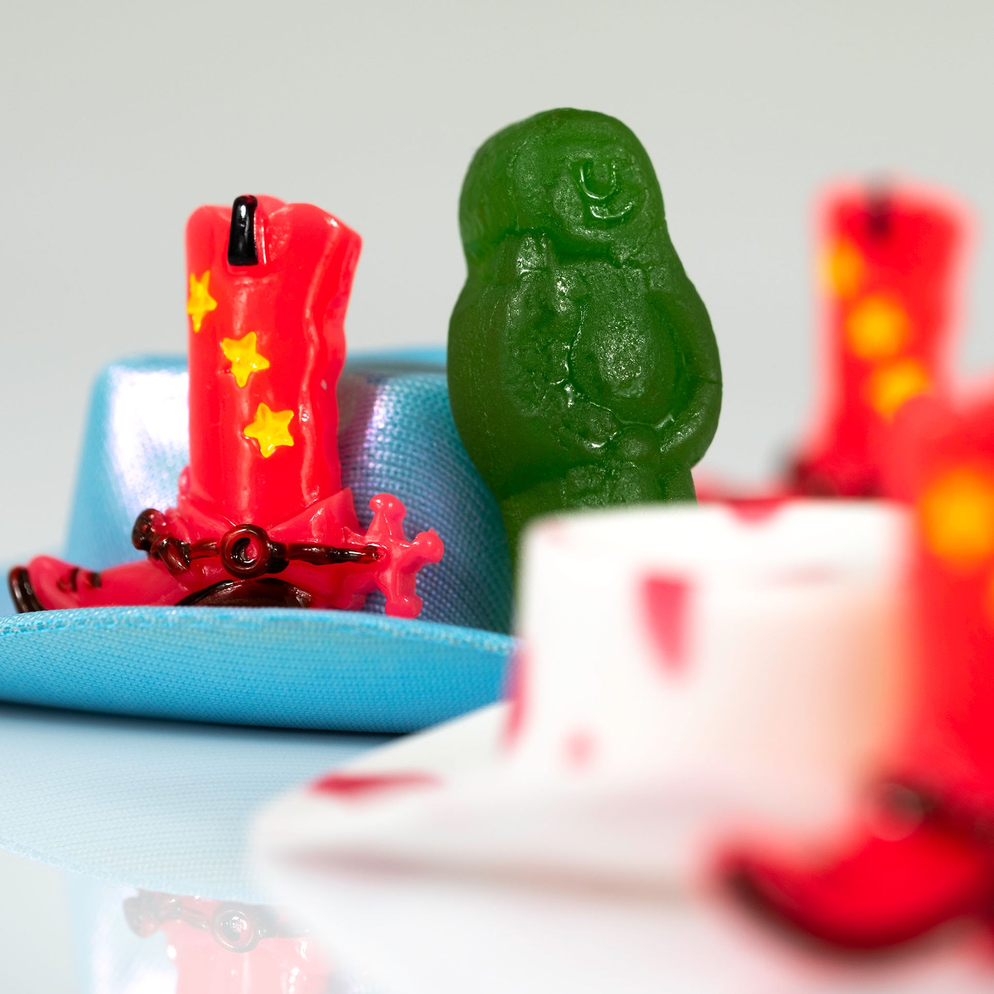 Square still life photograph of a red cowboy boot and green figure with turquoise, white and blurred red objects.