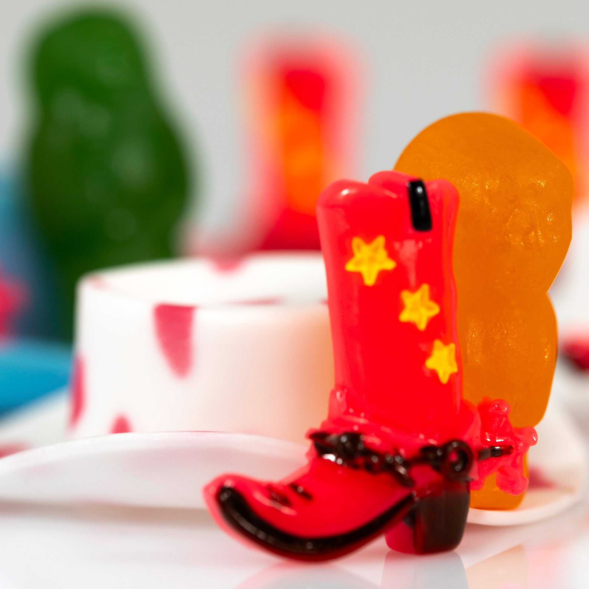 Close-up still life of a red cowboy boot with yellow stars beside an orange figure, with green and red objects blurred behind.