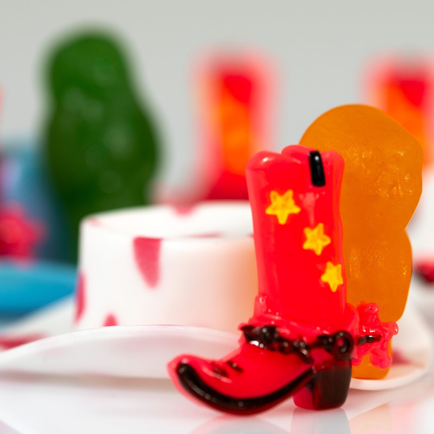 Square contemporary still life photograph featuring a red miniature cowboy boot, orange figure and layered green, white and red forms.