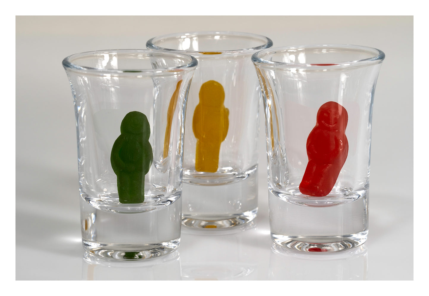 Three clear shot glasses containing green, yellow and red miniature figures arranged against a pale background.