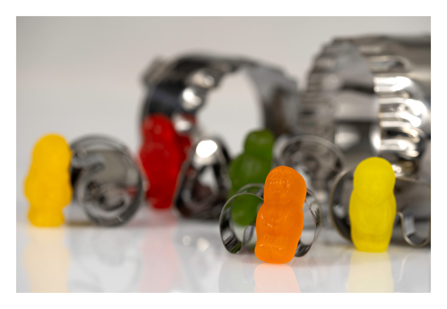 Colourful miniature figures arranged among scattered reflective metal baking cutters on a white surface.