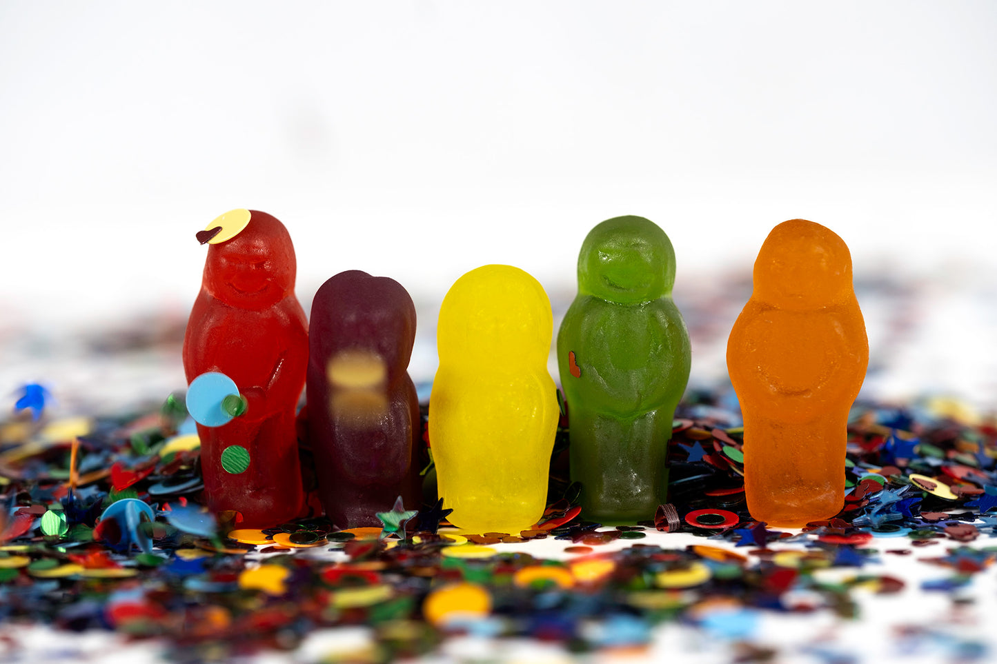 Five translucent red, purple, yellow, green and orange miniature figures arranged in a row amid scattered party glitter confetti.