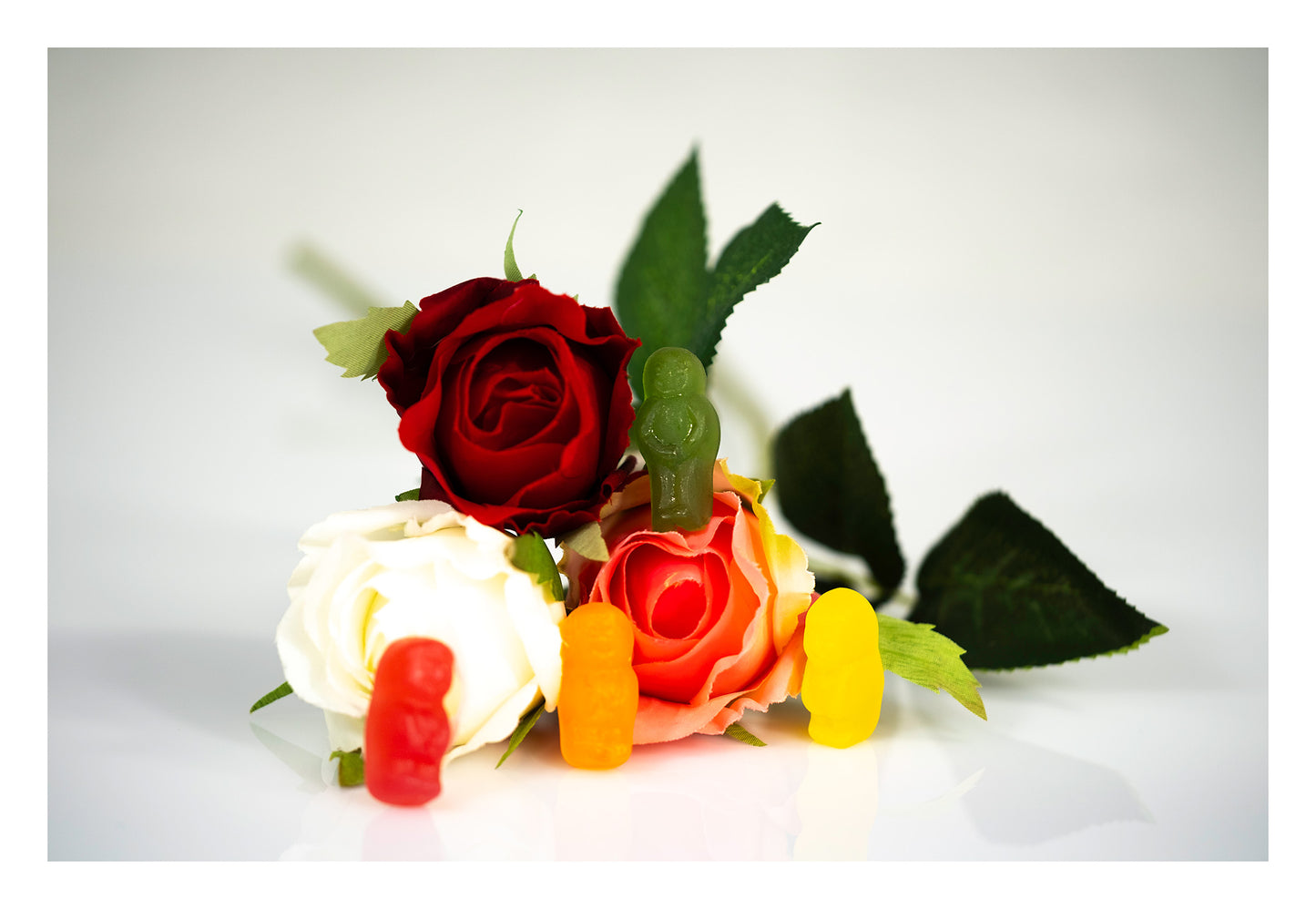 Red, orange, yellow and green gummy figures arranged among artificial red, white and pink roses with green foliage.