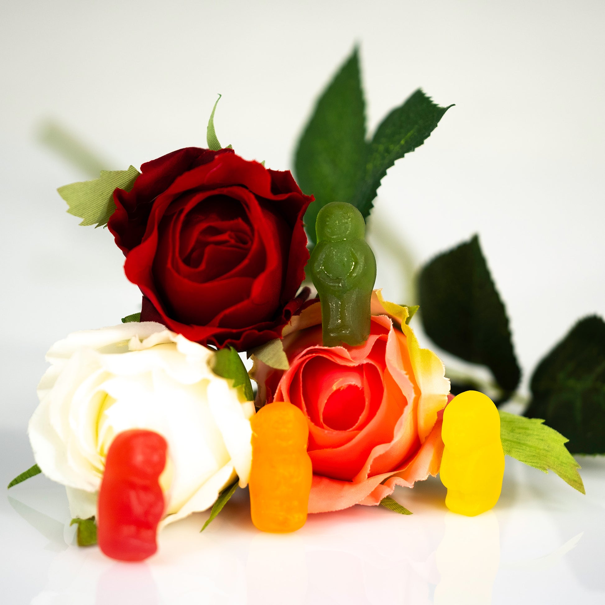 Close-up of four colourful gummy figures positioned around artificial red, white and pink roses against a pale background.