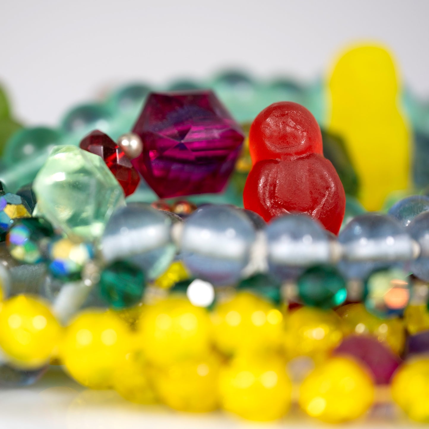 Close-up of a red gummy figure emerging among colourful beads and faceted jewellery with bright yellow forms blurred in the foreground.