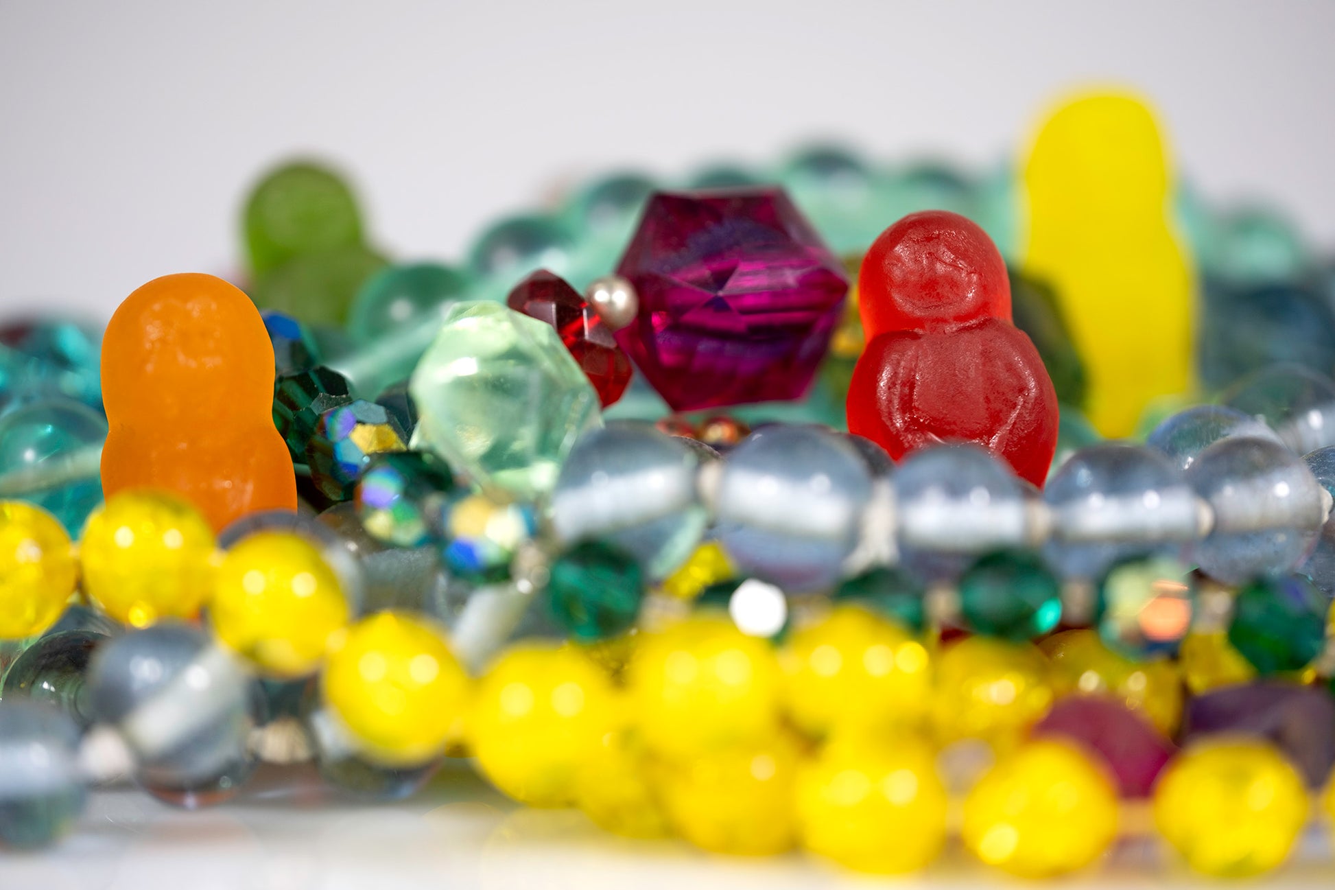 Orange, red, green and yellow gummy figures appearing among densely layered colourful beads and jewellery against a pale background.