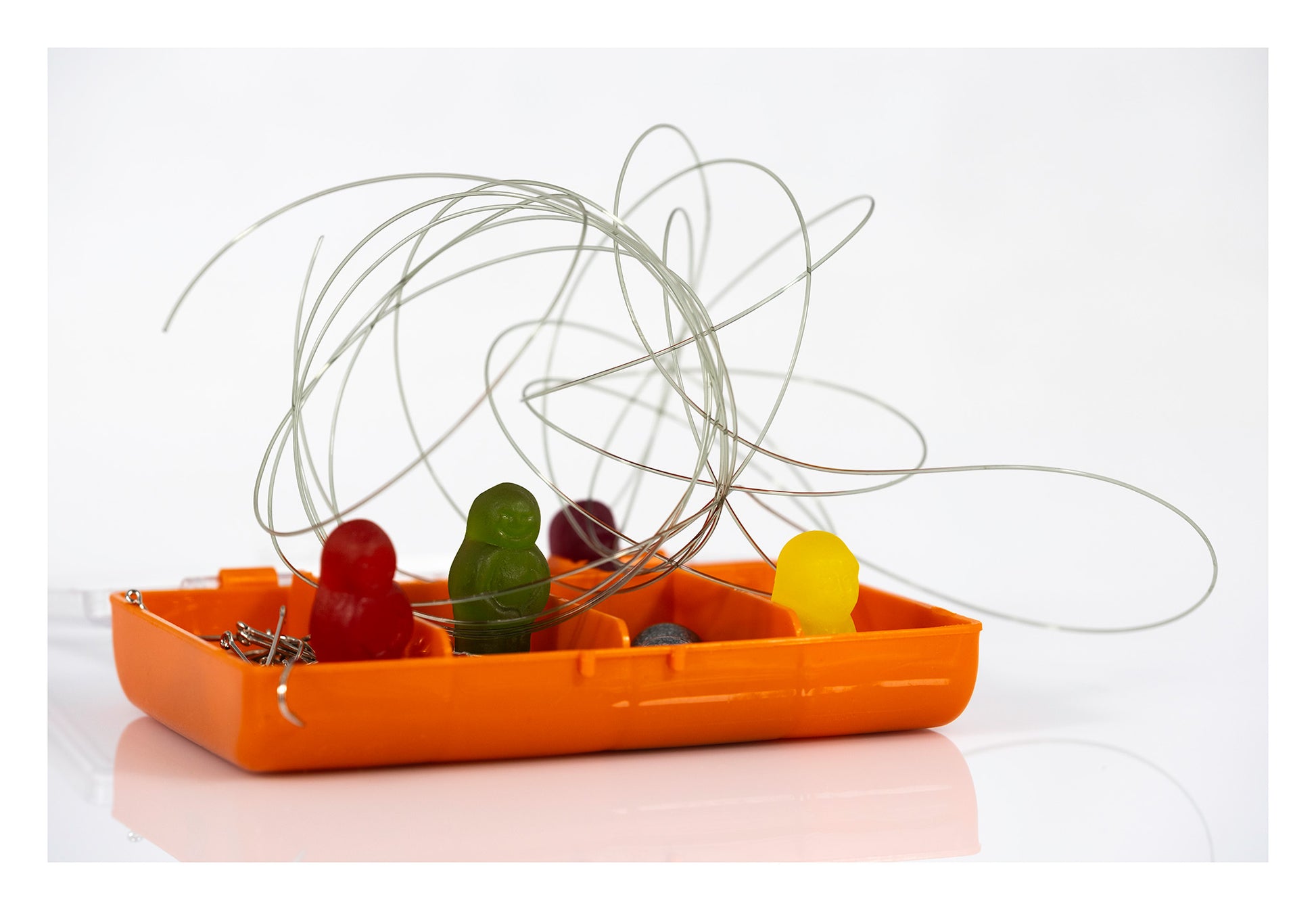Colourful gummy figures sitting inside an orange fishing tackle box surrounded by hooks, sinkers and looping fishing line.