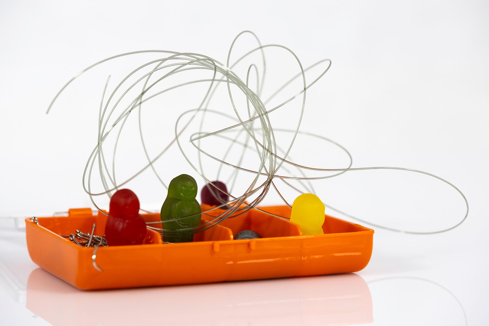 Orange fishing tackle box containing colourful gummy figures, hooks and sinkers beneath large looping strands of fishing line against white.