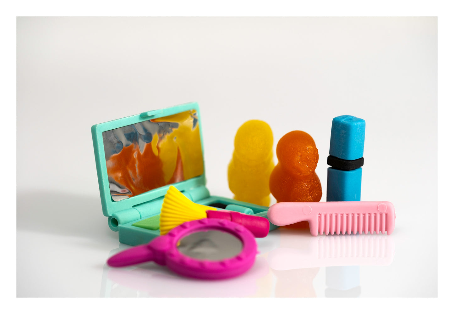 Yellow and orange gummy figures surrounded by colourful miniature mirrors, a comb, brush and lipstick on a white background.