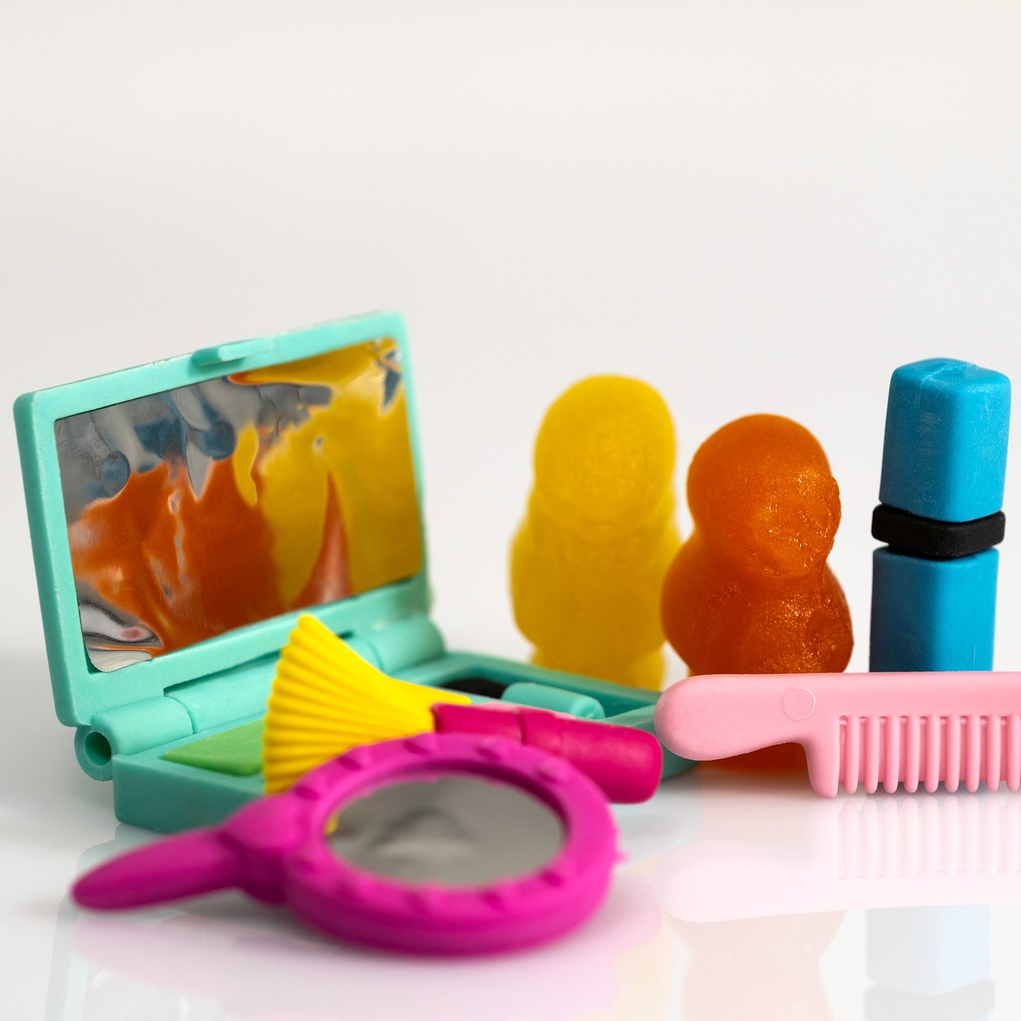 Close view of yellow and orange gummy figures beside a turquoise compact mirror, blue lipstick and pink comb, with a magenta hand mirror in the foreground.