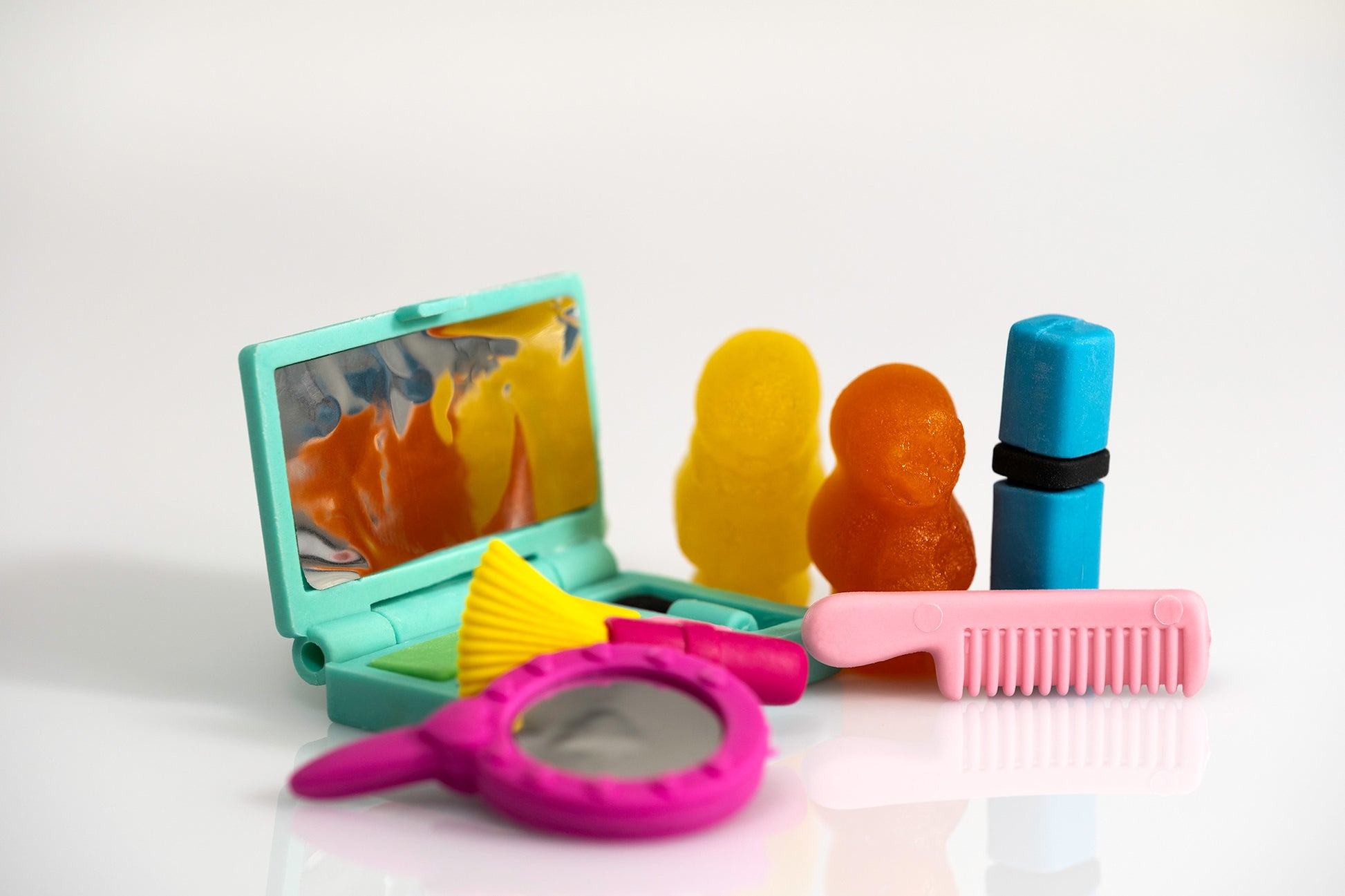 Colourful gummy figures with miniature beauty accessories including a turquoise compact mirror, pink comb, brush, hand mirror and blue lipstick.