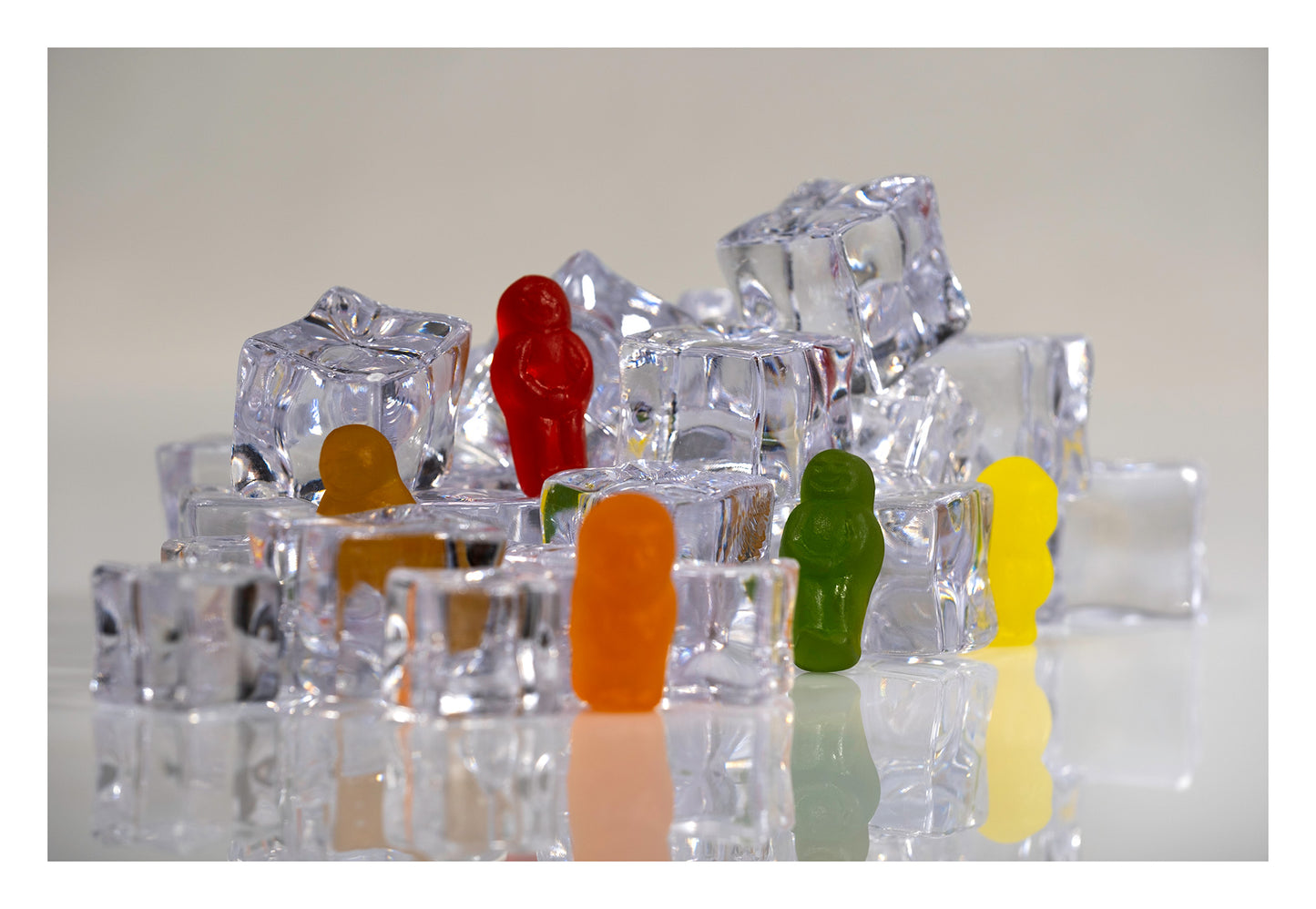 Red, orange, green and yellow gummy figures arranged among stacked transparent ice cubes on a reflective white surface.