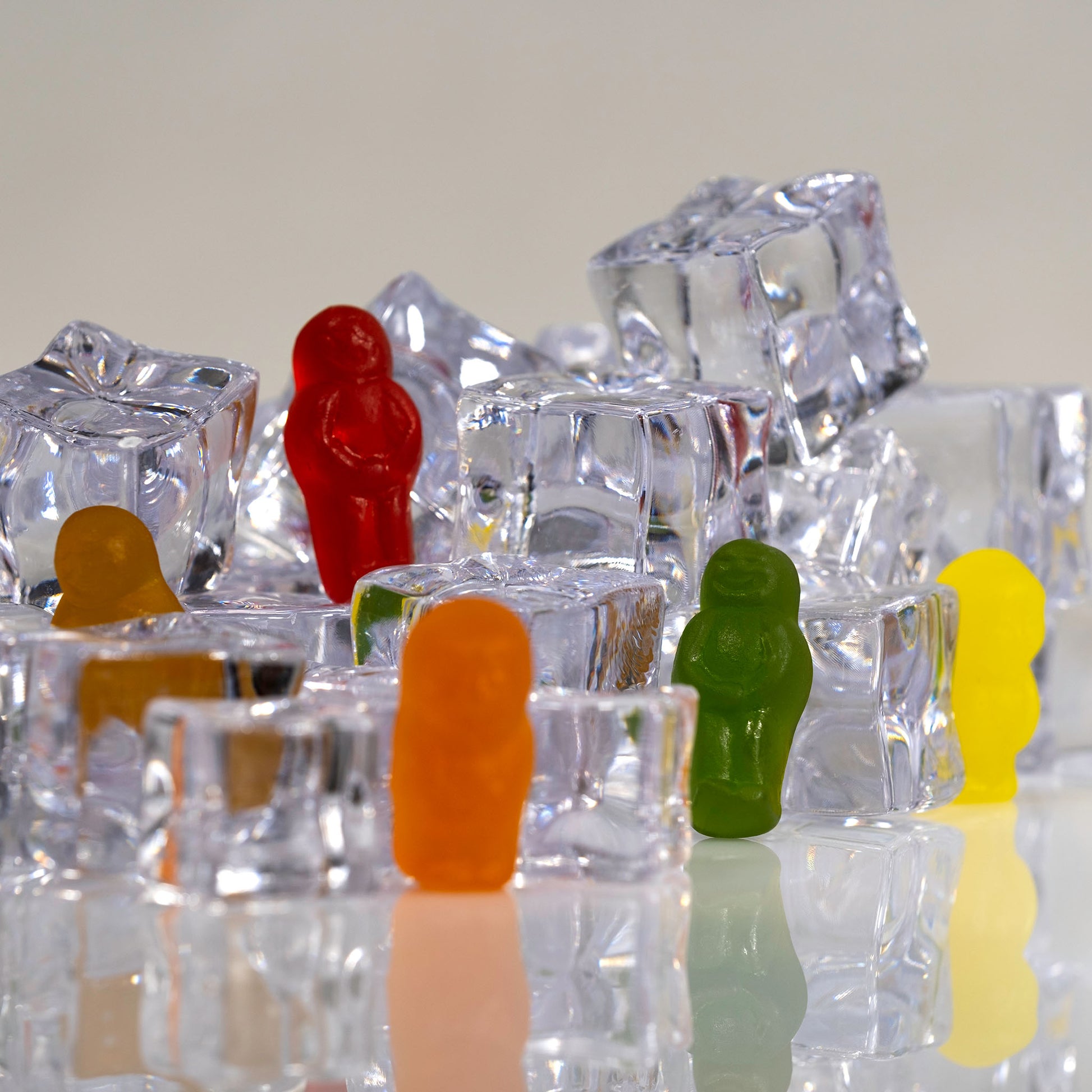 Close square view of colourful gummy figures standing among transparent ice cubes with reflections on a glossy surface.