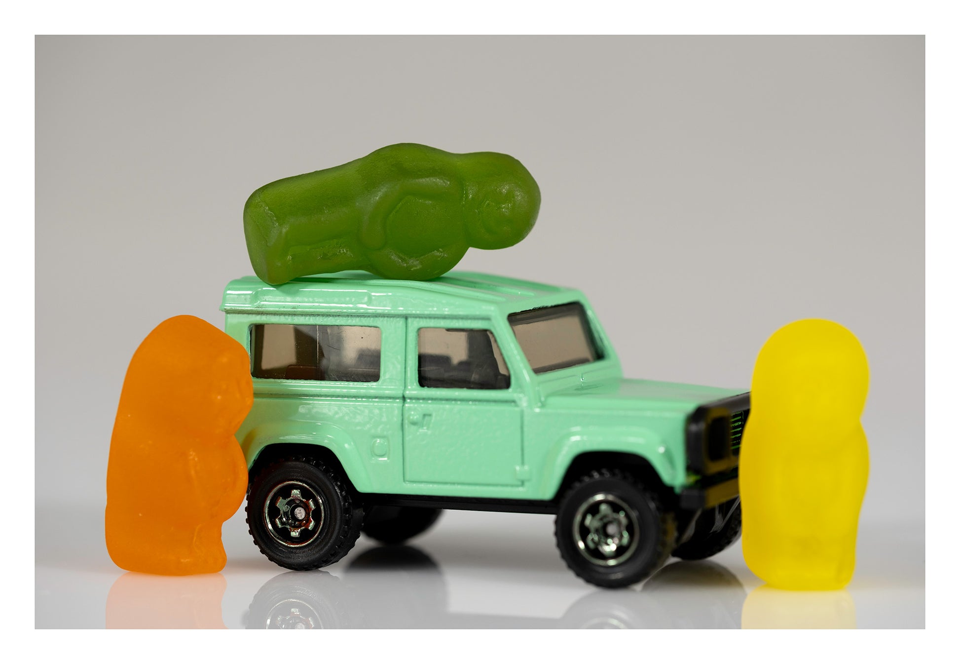 Orange and yellow gummy figures standing on opposite sides of a mint-green miniature four-wheel drive with a green gummy figure lying across its roof.