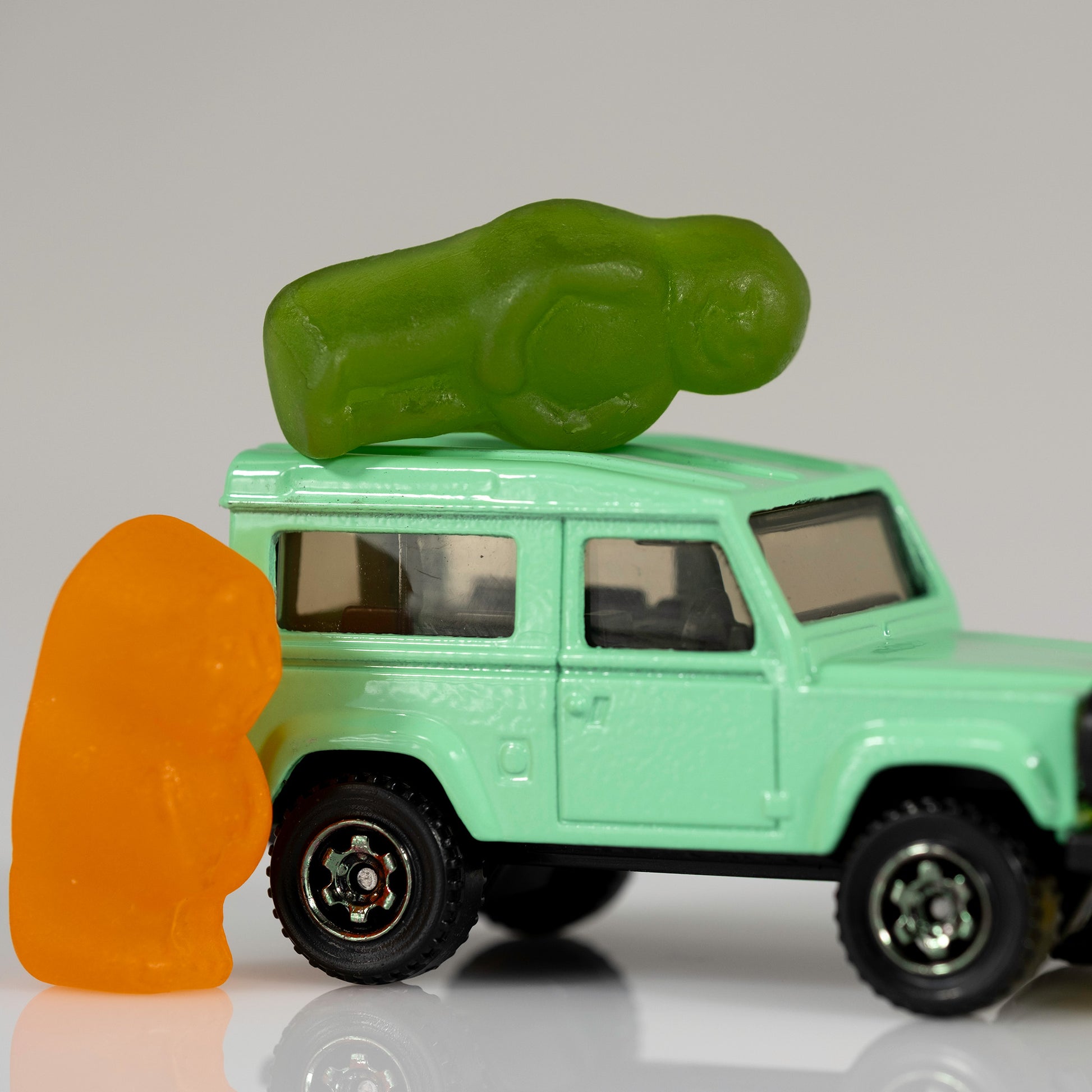 Close square crop of an orange gummy figure beside a mint-green toy four-wheel drive with a green gummy figure lying on the roof.
