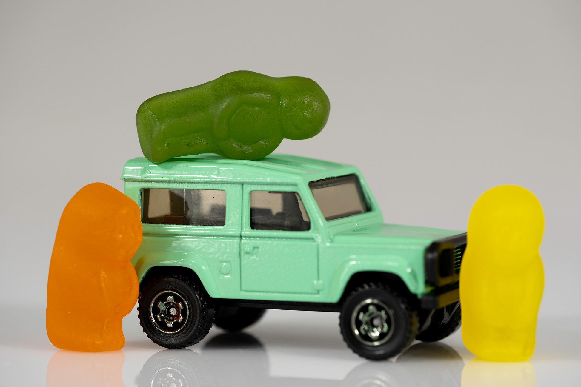 Orange and yellow gummy figures flanking a mint-green miniature four-wheel drive while a green gummy figure lies horizontally across the roof.