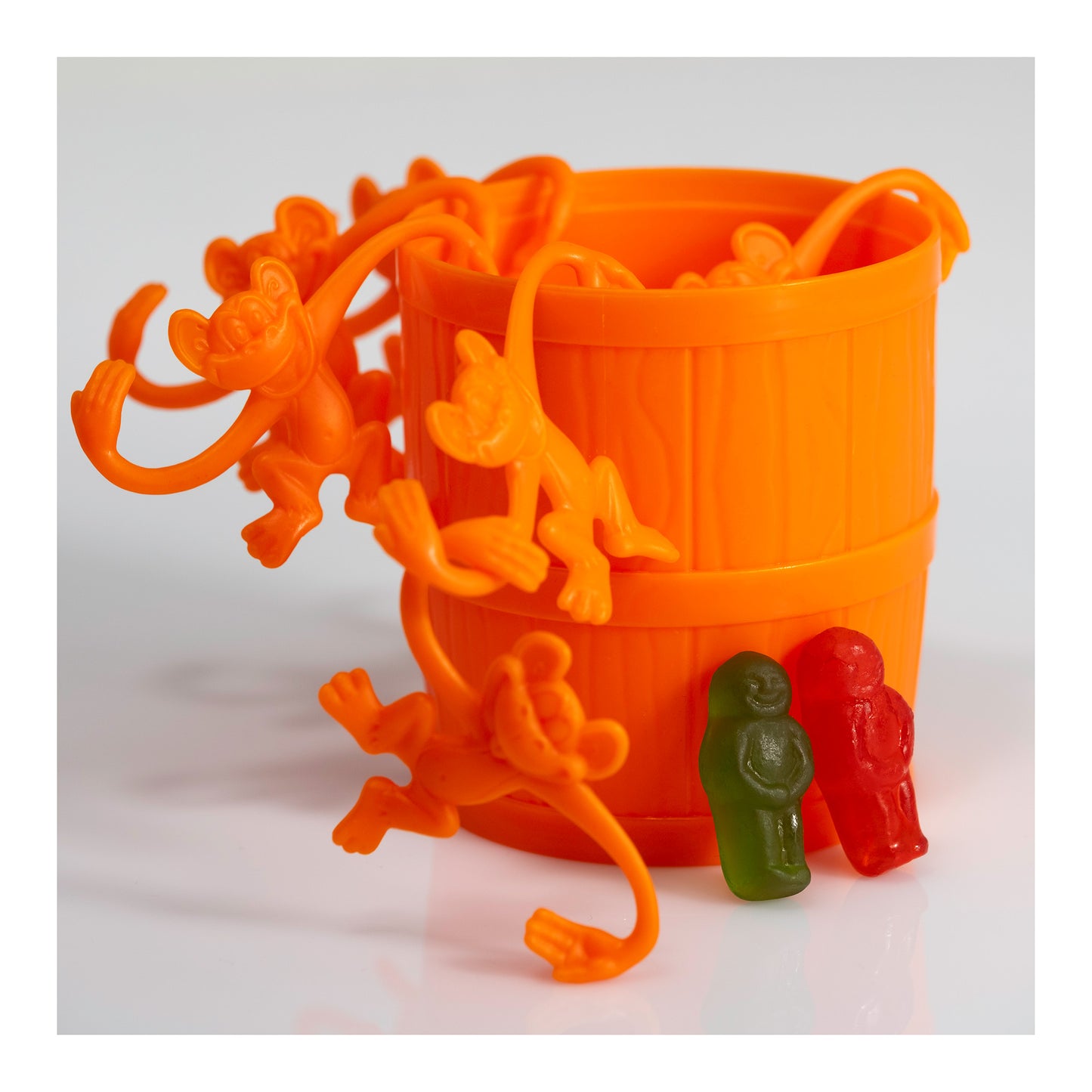 Orange toy monkeys hanging from and climbing around an orange barrel beside green and red gummy figures on a white reflective surface.