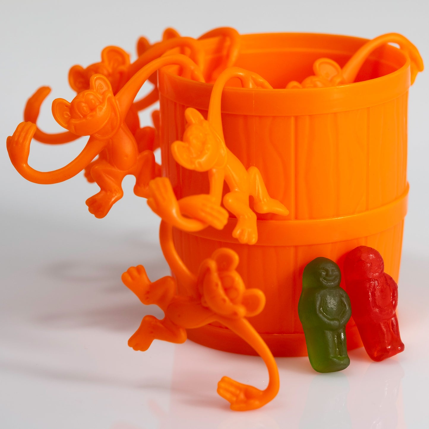 Close view of bright orange toy monkeys clustered around an orange barrel with green and red gummy figures standing at the lower right.