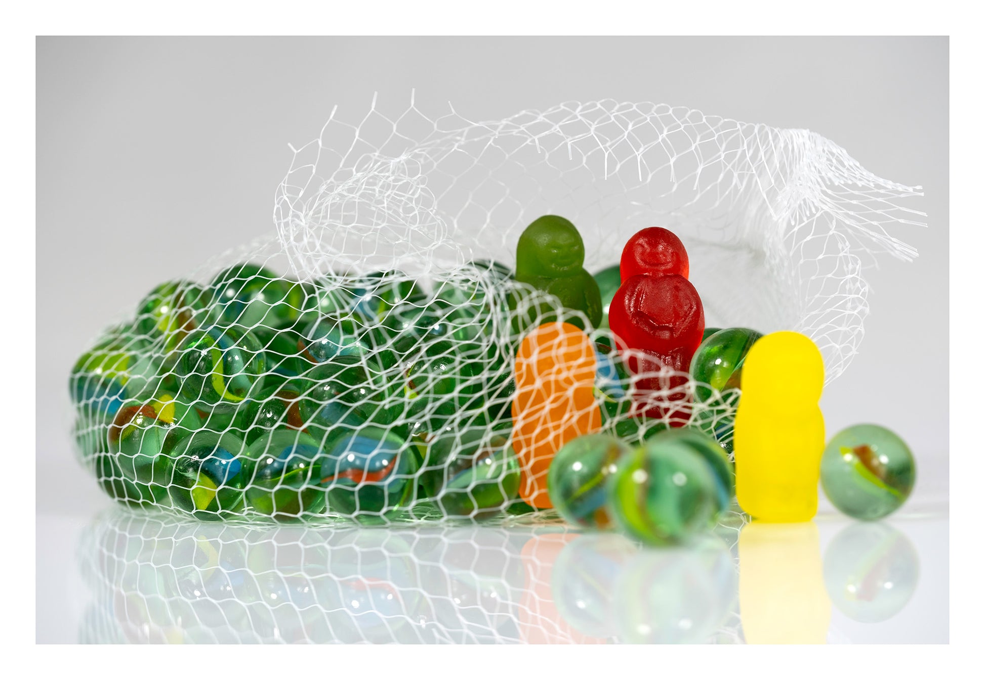 Glass marbles spilling from a white mesh bag among green, red, orange and yellow gummy figures on a reflective white surface.