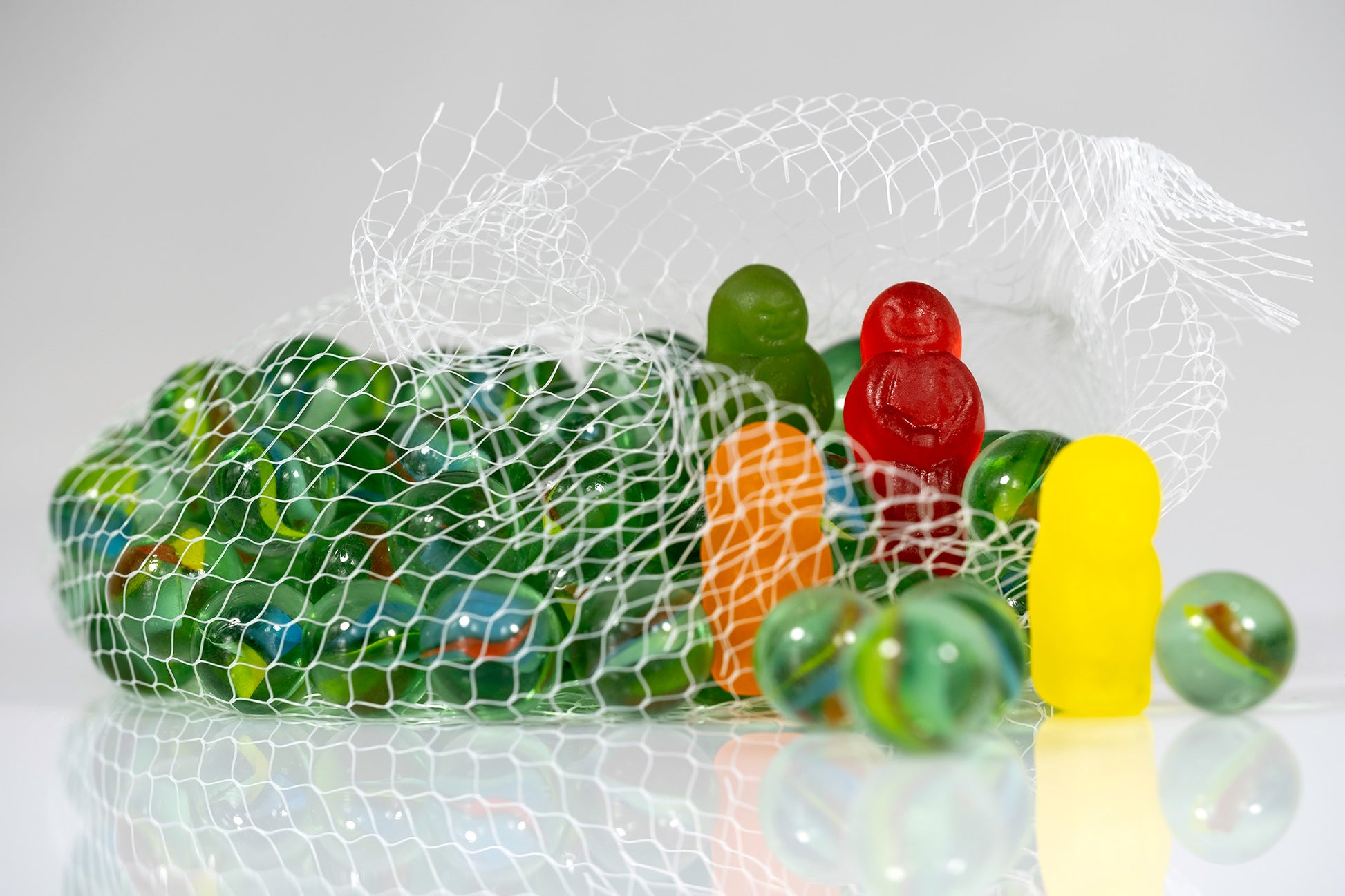 Green glass marbles spilling from an open mesh bag surrounded by colourful gummy figures and reflected on a white surface.