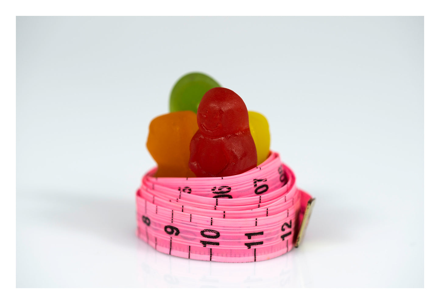 Red, orange, green and yellow gummy figures gathered inside a coiled pink measuring tape against a white background.