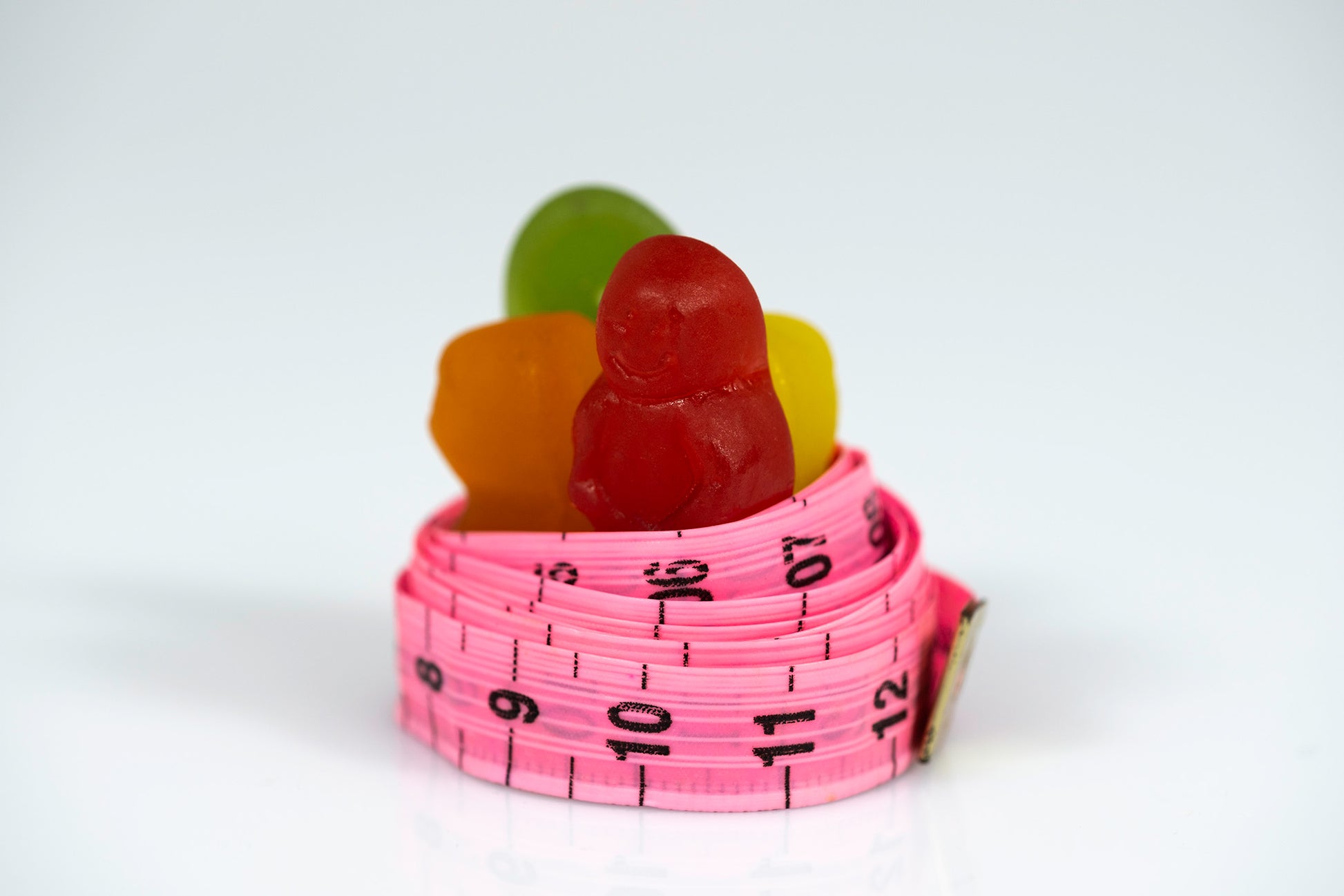 Colourful gummy figures emerging from a coiled pink measuring tape on a white reflective surface.