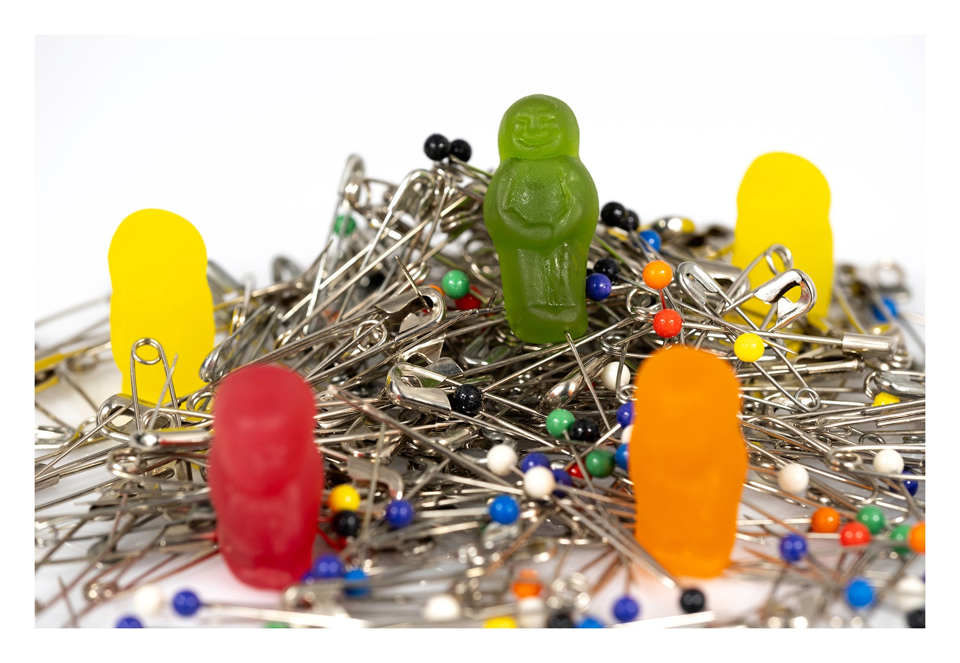 Colourful gummy figures arranged among a large tangled pile of silver safety pins and multicoloured dressmaking pins against a white background.