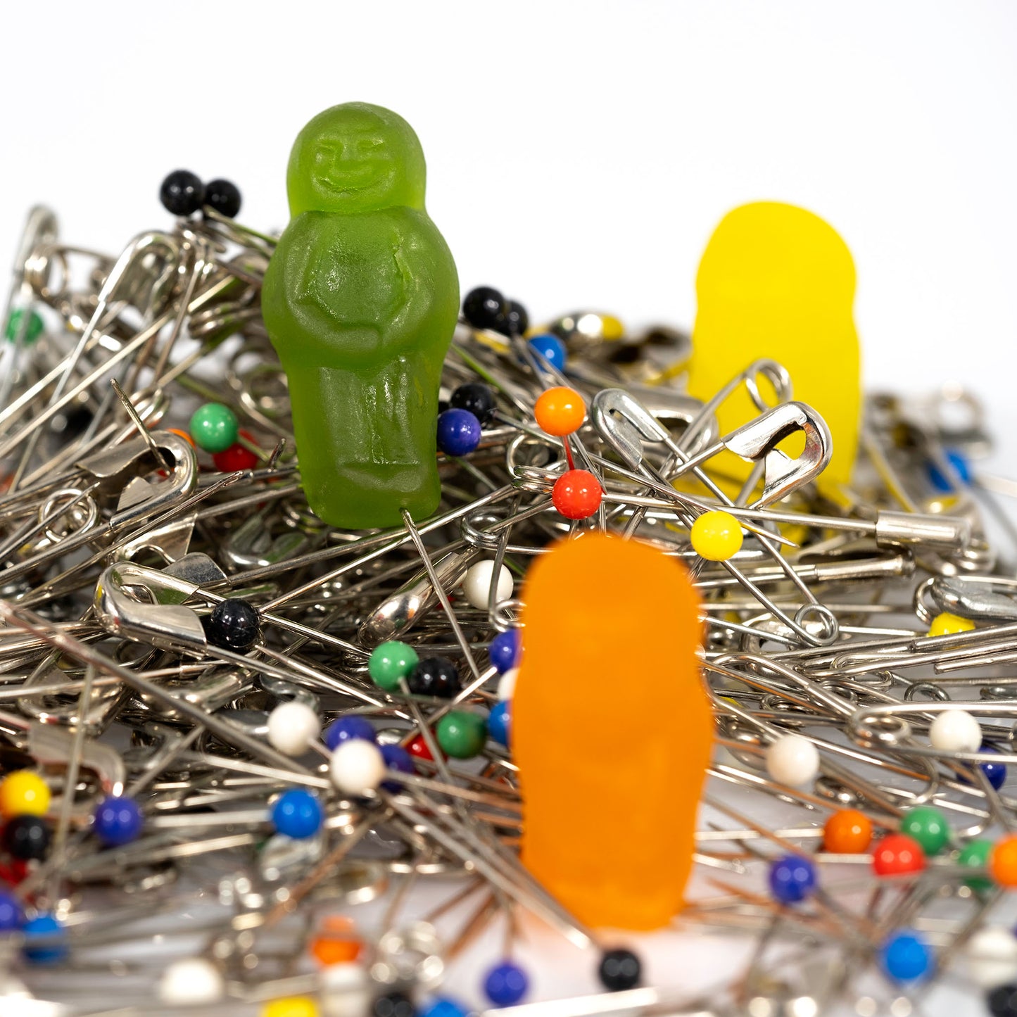 Close-up of a green gummy figure standing among silver safety pins and coloured dressmaking pins, with an orange figure blurred in the foreground.