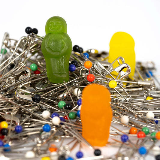 Close-up of a green gummy figure standing among silver safety pins and coloured dressmaking pins, with an orange figure blurred in the foreground.
