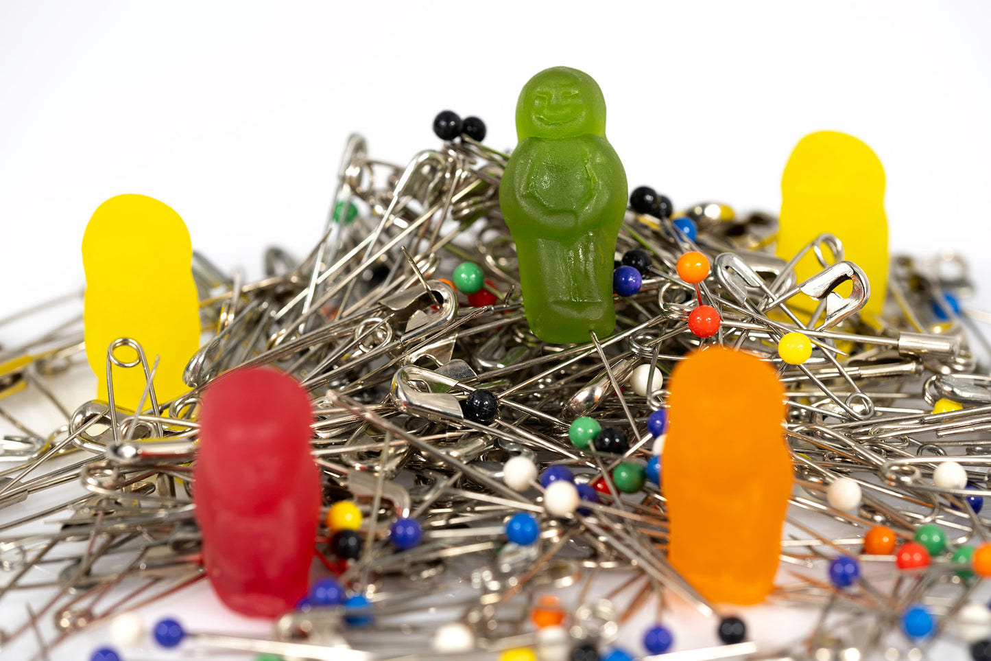 Green, red, orange and yellow gummy figures positioned throughout a tangled pile of silver safety pins and multicoloured dressmaking pins.